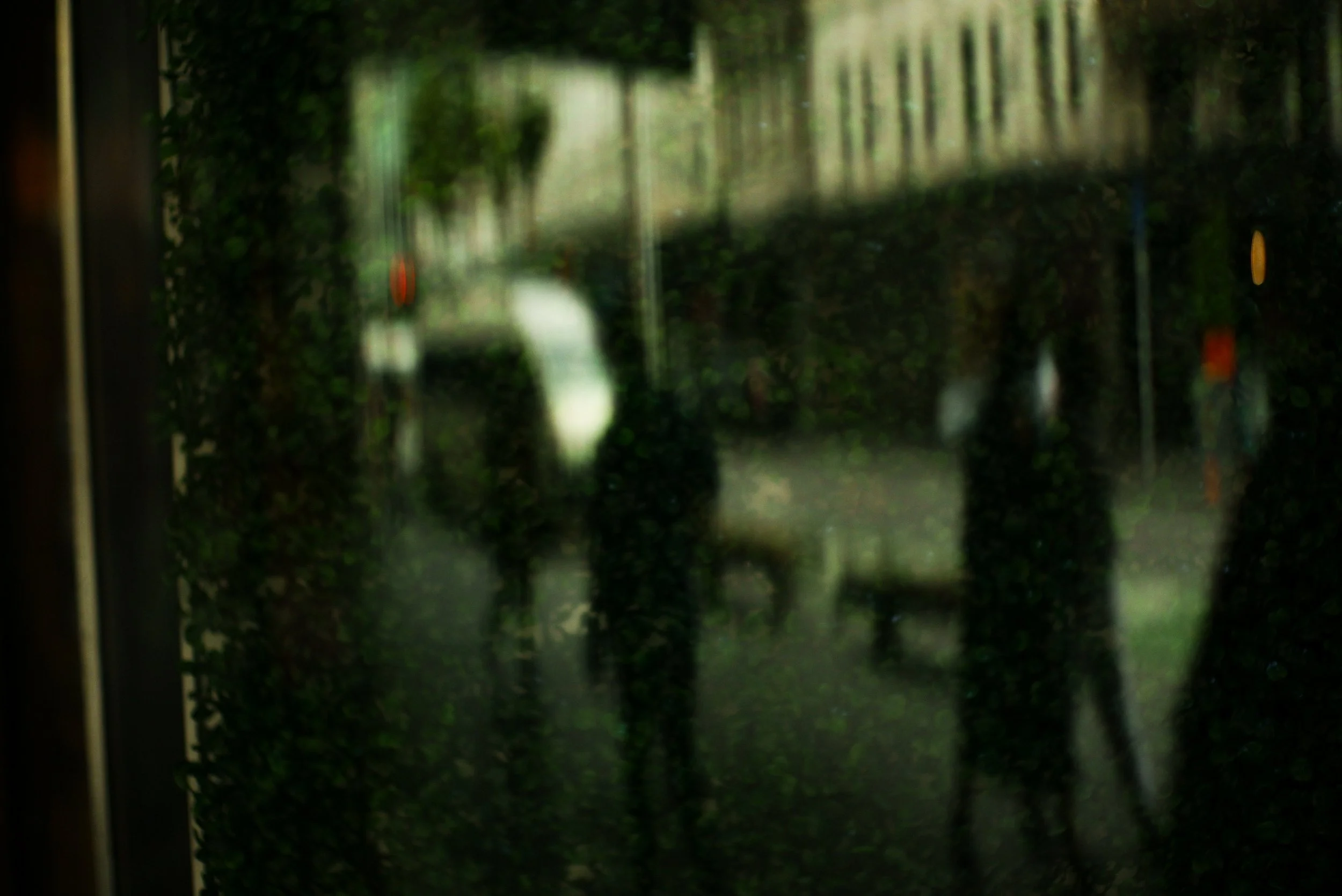Blurry view of people walking on a sidewalk through a glass window or door, with trees and a vehicle in the background.