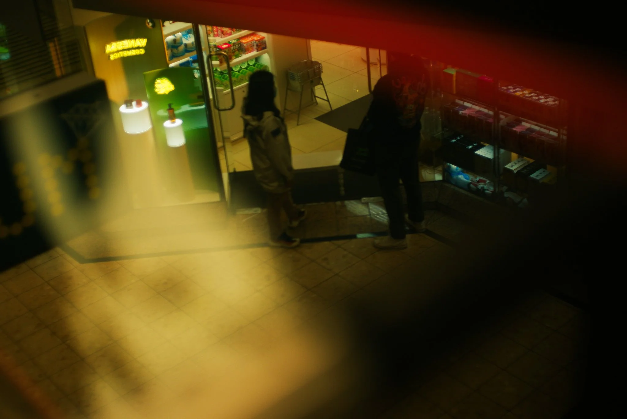 Two people standing at the entrance of a store, viewed from above through a semi-opaque surface. The store has shelves with various products and bright lighting.