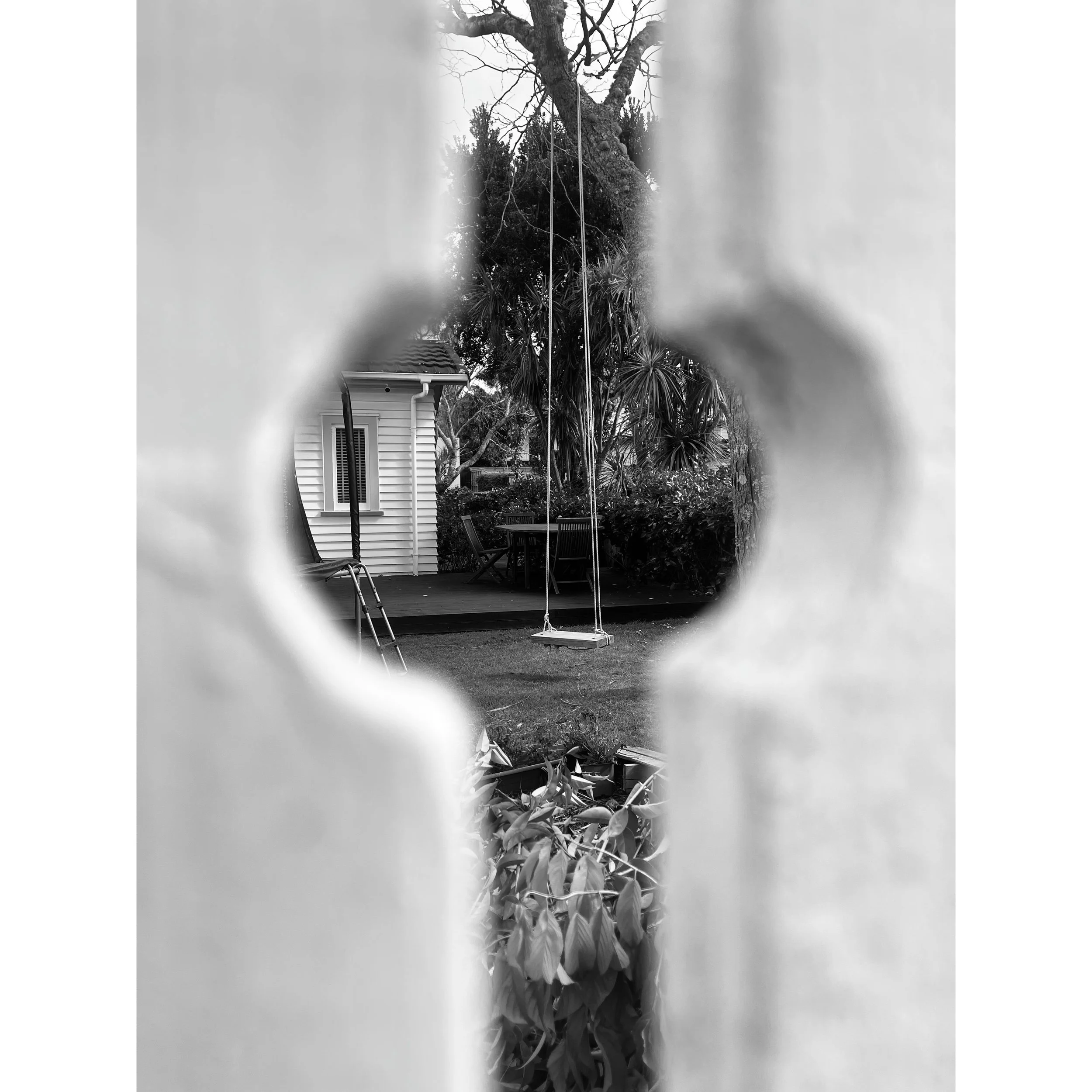View through a cutout shape in a wall to a backyard with a swing set, table, chairs, trees, and bushes in black and white