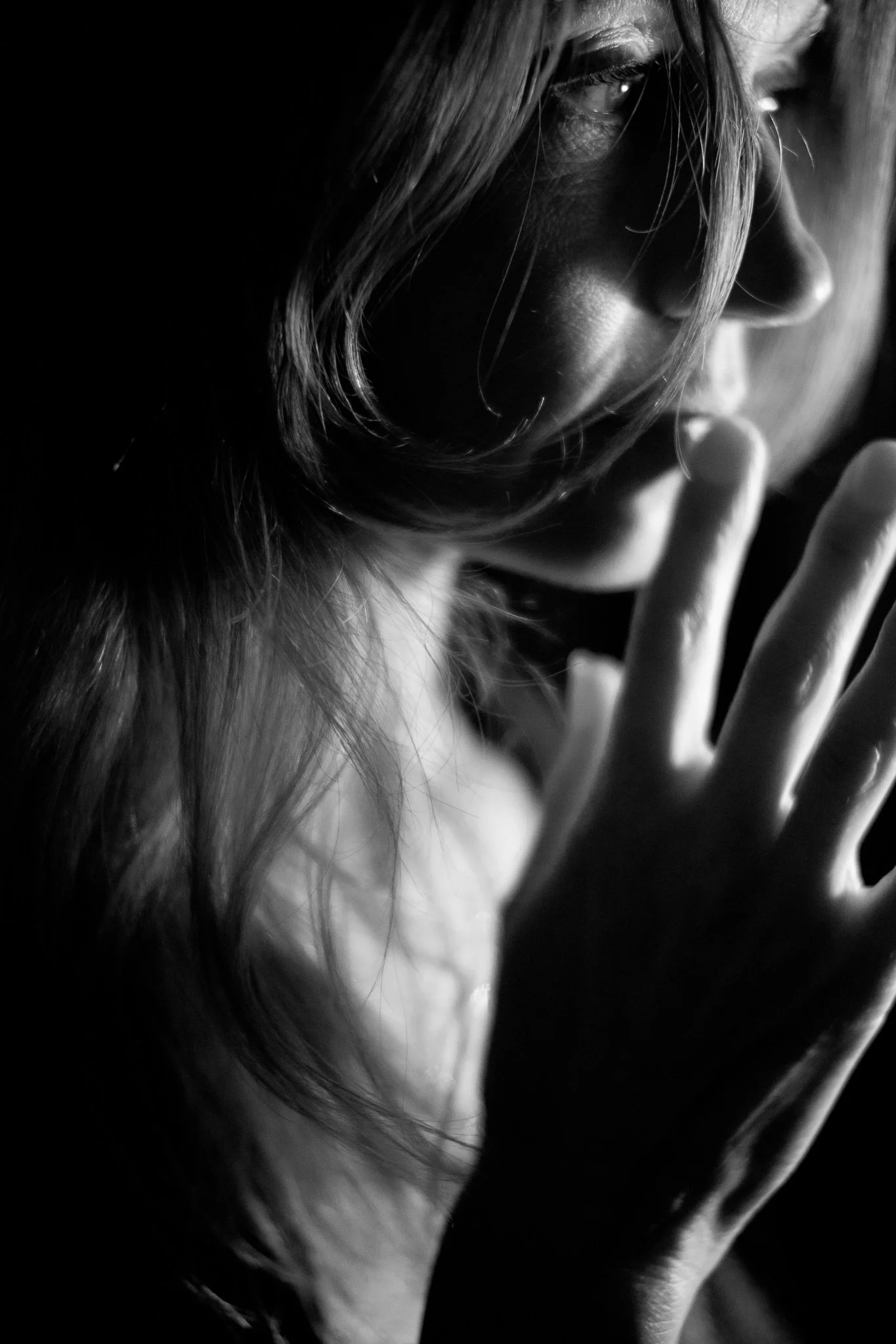 Close-up black and white photo of a woman with long hair, partially covering her face, holding her finger near her lips in a shushing gesture.