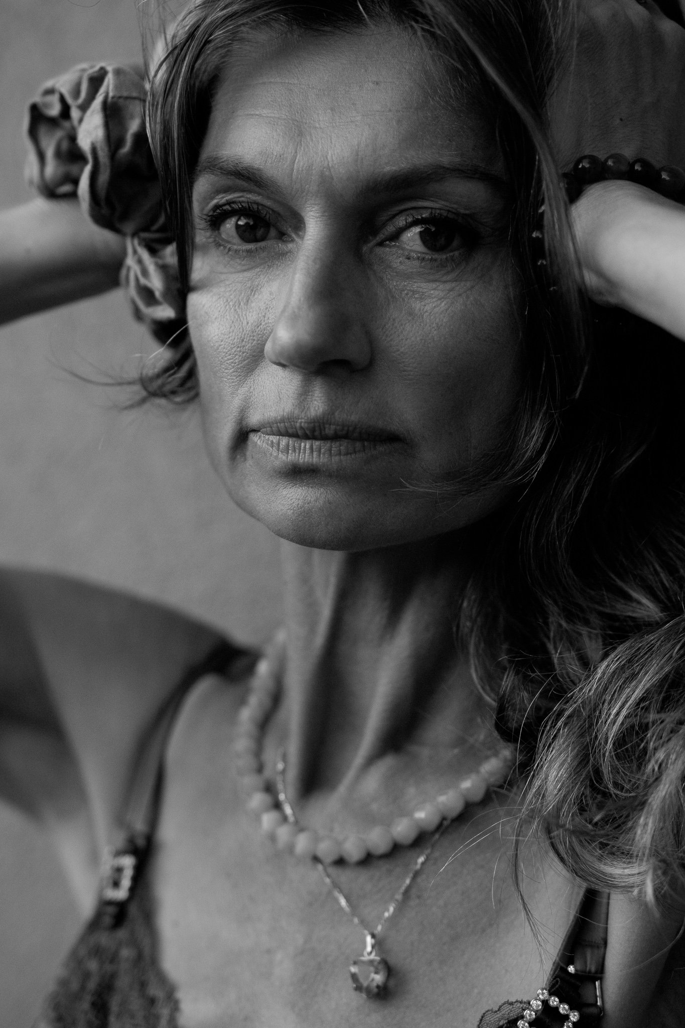 A black-and-white close-up portrait of a woman with wavy hair. She has a serious expression, and is wearing necklaces and a black strap top. She is lying down with one arm behind her head and a beaded bracelet on her wrist.