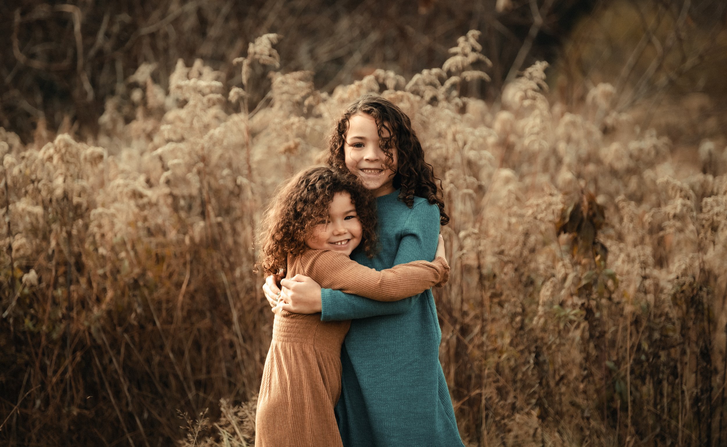Two young girls with curly hair hugging each other in a field of tall, dry grass during autumn.