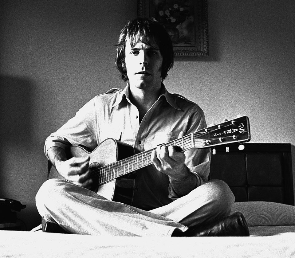 A man sitting cross-legged on the bed and playing an acoustic guitar while looking at the camera