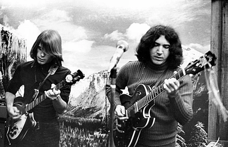 Two men playing guitars in front of a mural of mountains