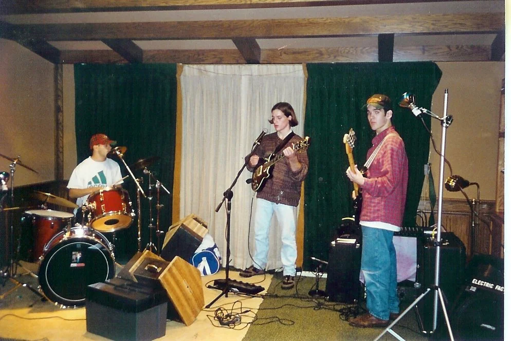 Three young men playing drums, guitar, and bass at a house party