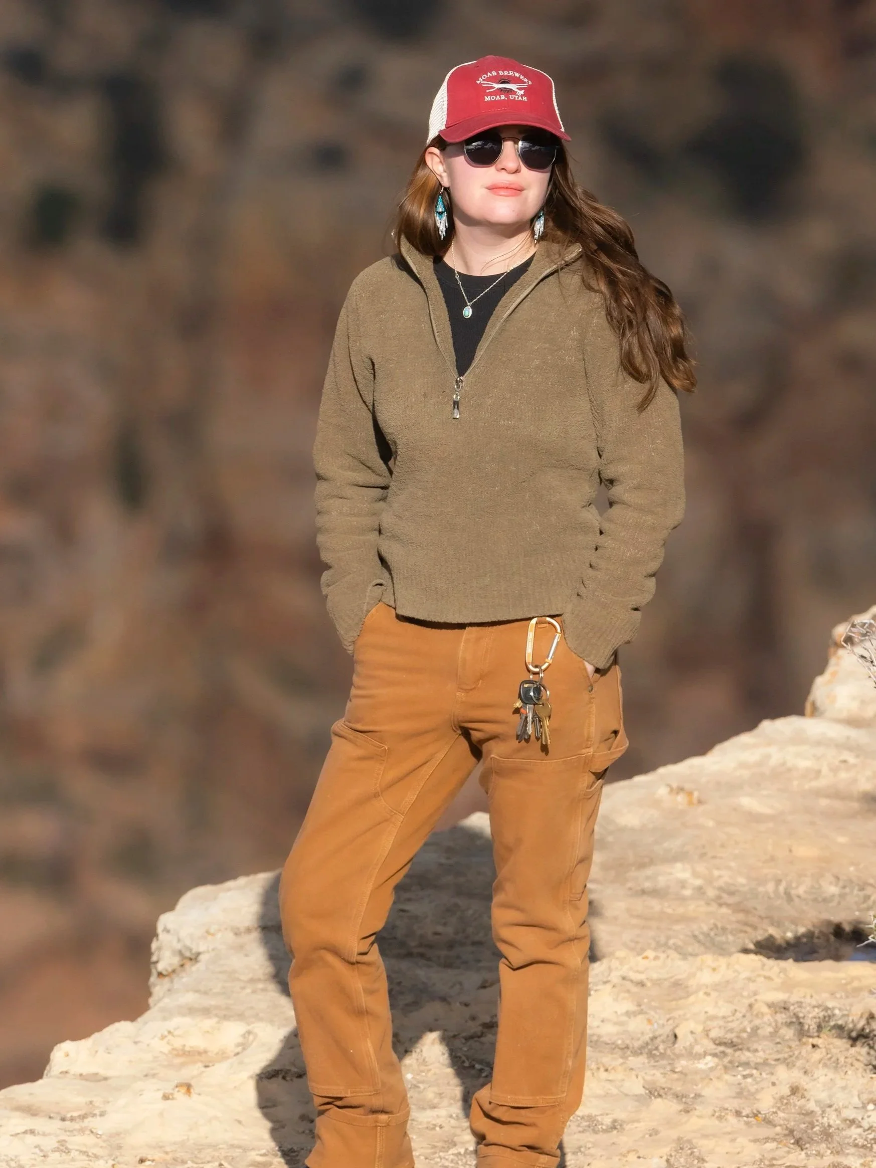 Woman standing outdoors on rocky terrain, wearing sunglasses, a red and white cap, a brown fleece jacket, and brown pants with a set of keys hanging from her pocket.