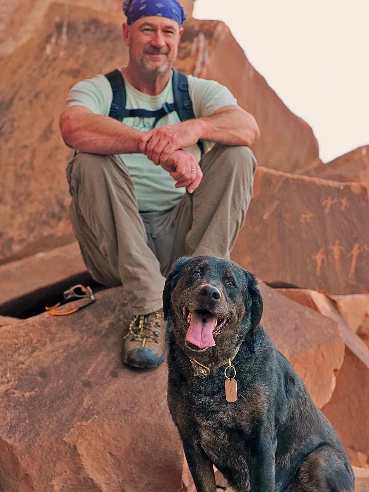 A man in outdoor clothing with a blue bandana seated on a rocky terrain next to a happy black dog with a collar, both surrounded by reddish rocks.