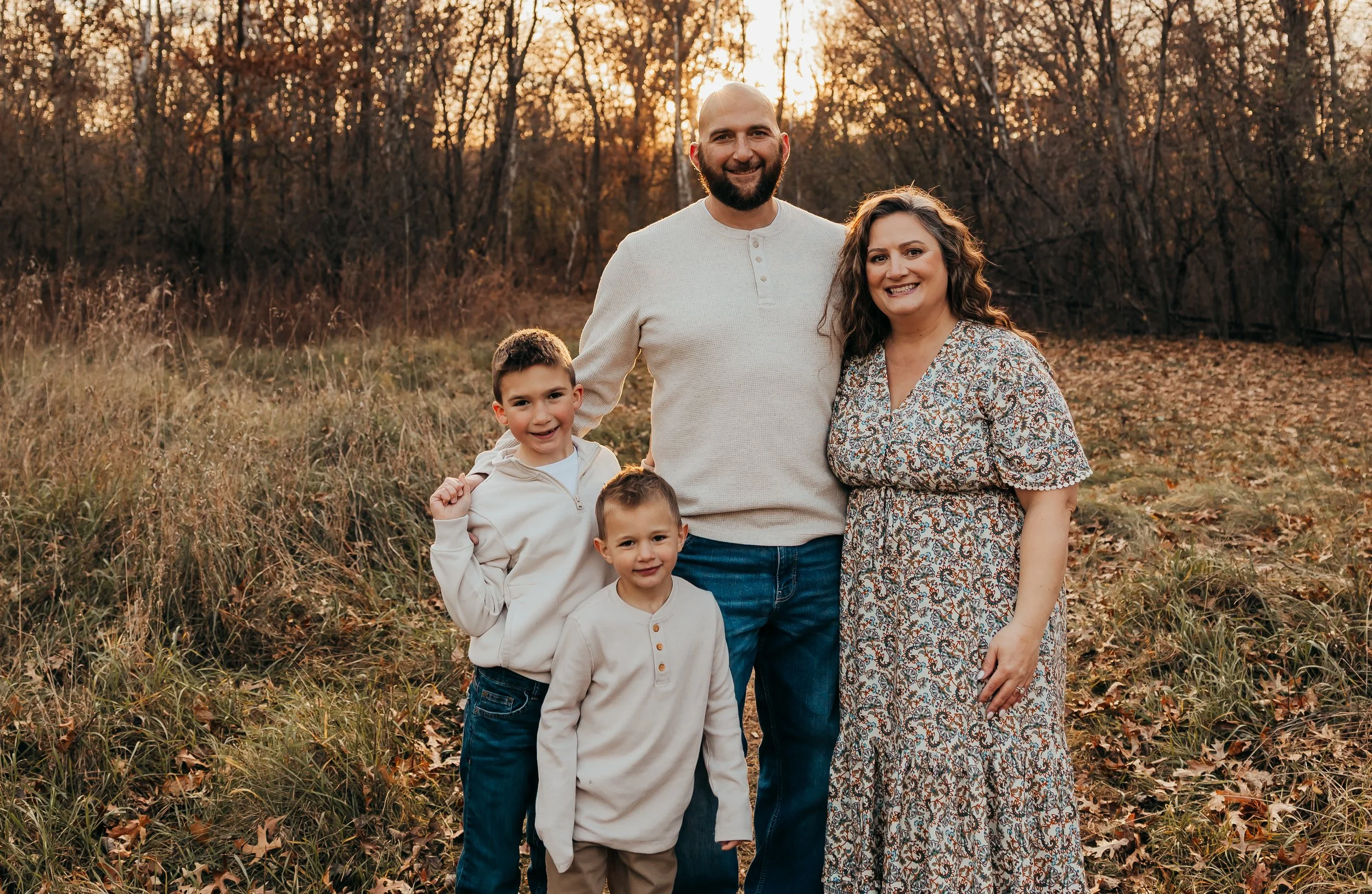 Family of five smiling outdoors during autumn, standing on fallen leaves with trees and sunset in the background.
