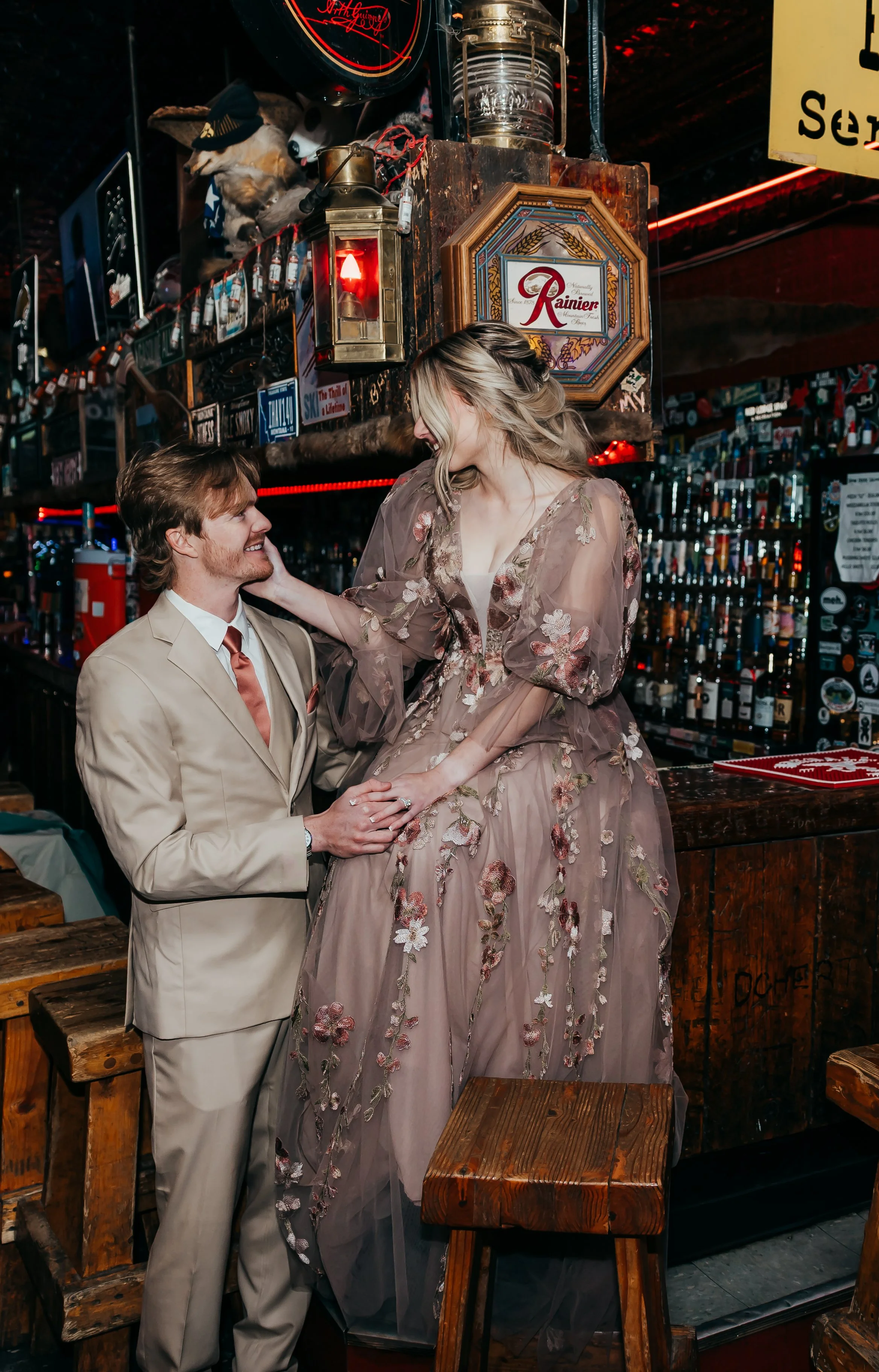 A couple is celebrating at a bar, with the man in a beige suit and the woman in a sheer floral dress. They are smiling and holding hands, with the woman sitting on the bar counter.