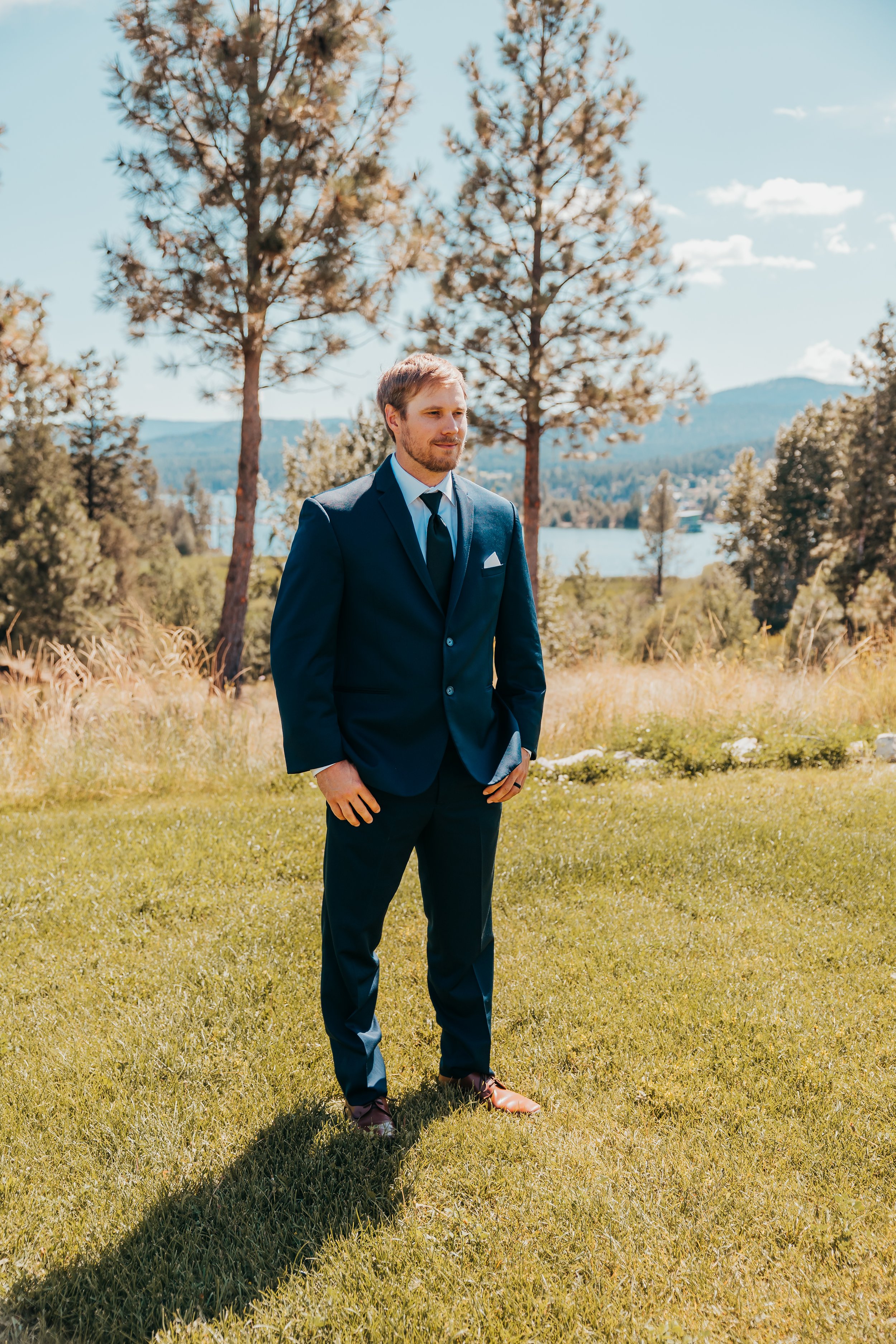 A man in a navy blue suit and tie standing outdoors on a grassy area with trees and mountains in the background.