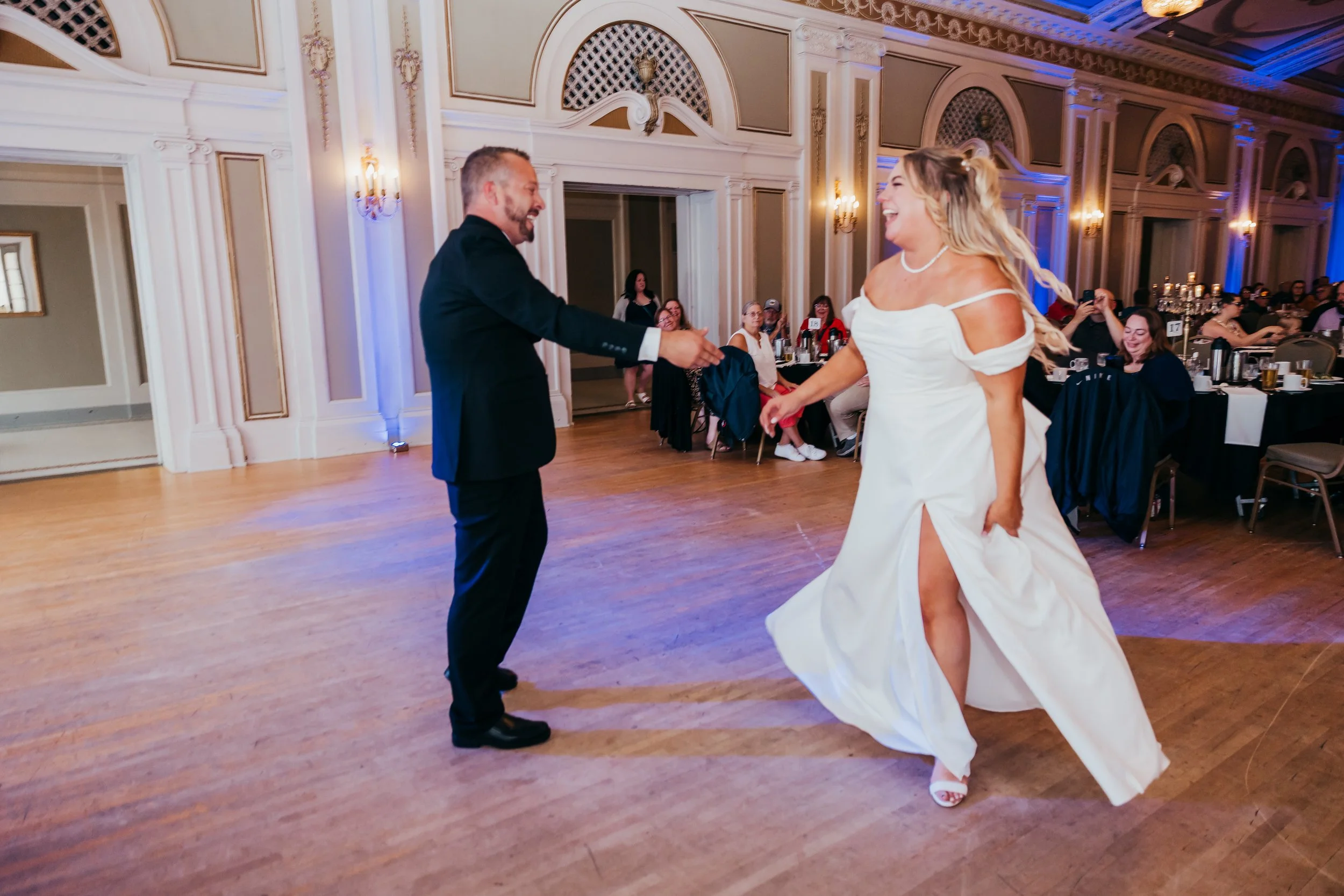 A bride and groom dancing and smiling at their wedding reception, with guests seated at tables in the background.