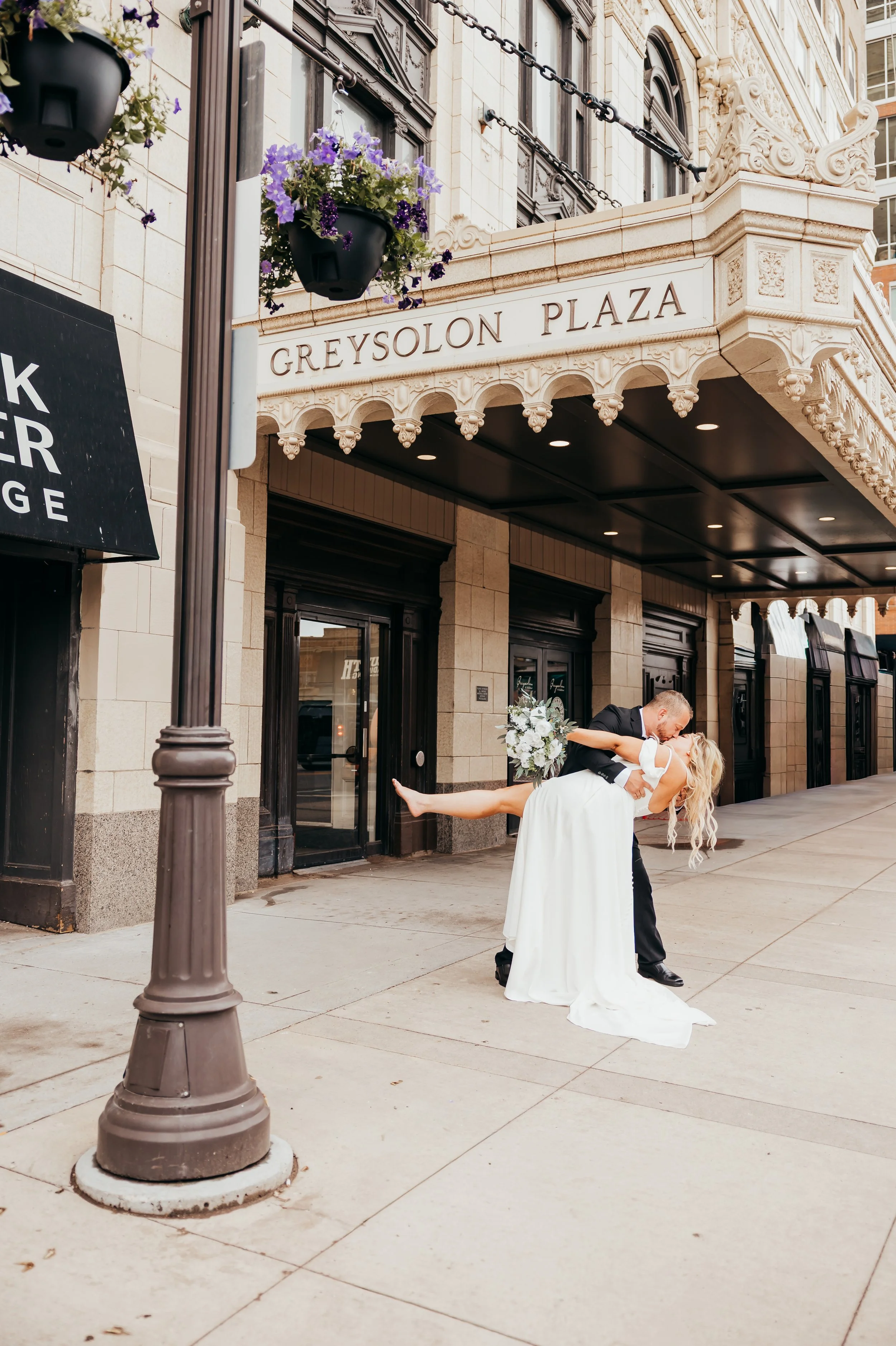 A newlywed couple sharing a kiss outside Greysolon Plaza, the groom in a black tuxedo dipping the bride in a white wedding dress, holding a bouquet of flowers, in front of a historic building with ornate architecture and hanging flower baskets.