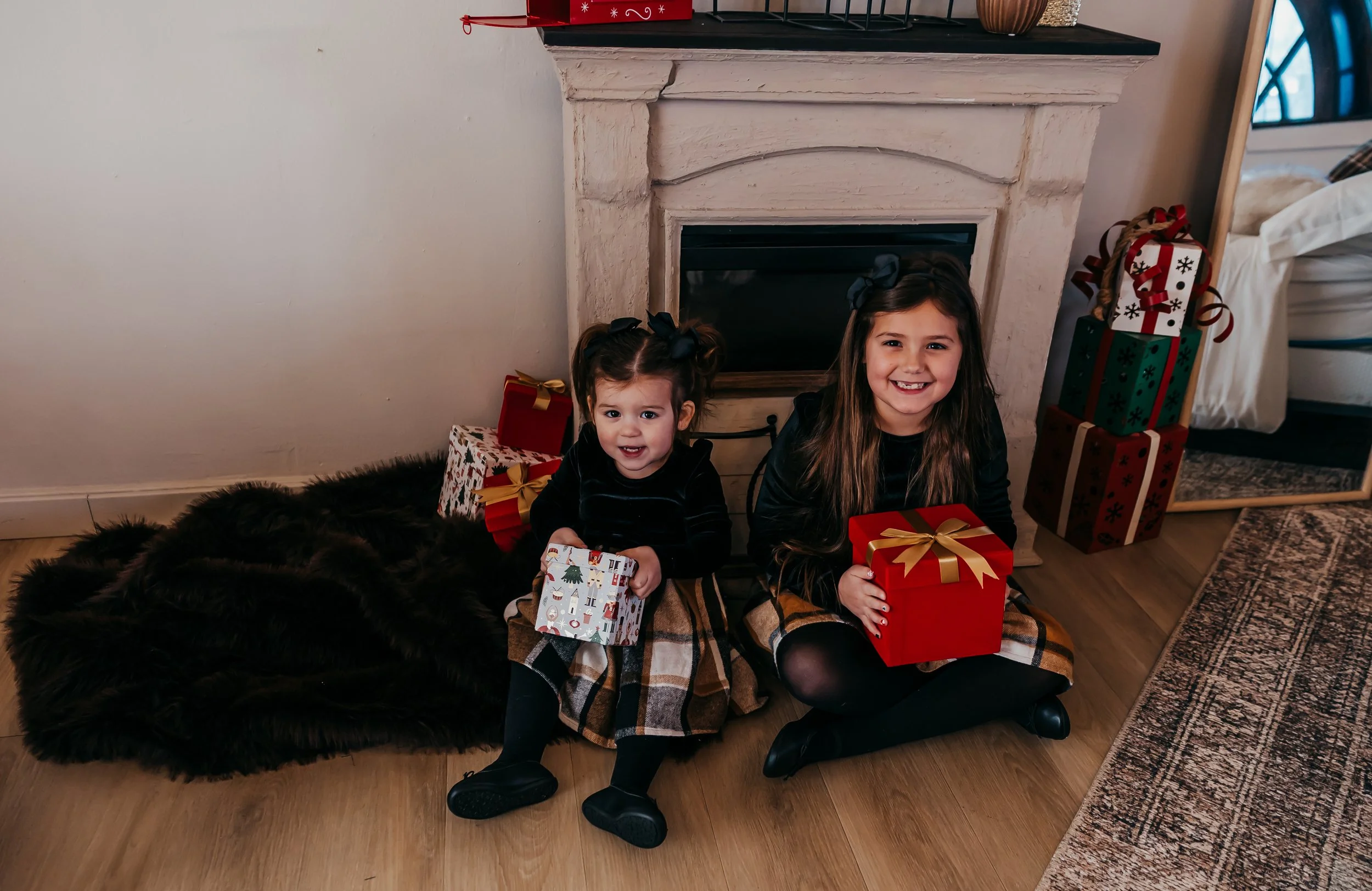 Two young girls sitting in front of a fireplace holding Christmas presents, with wrapped gifts stacked nearby.