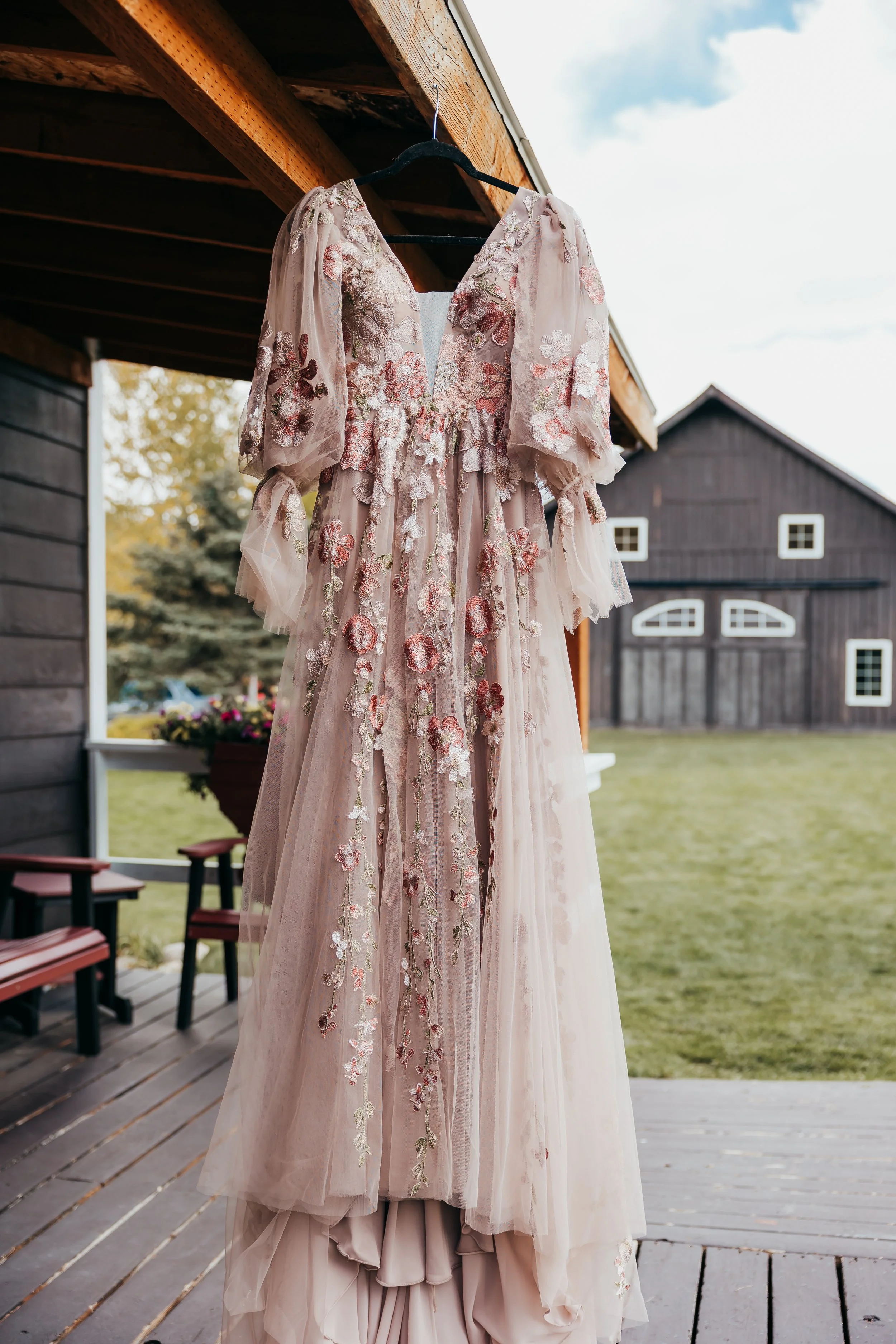 A pink embroidered gown hanging on a black hanger outside a rustic barn on a porch with wooden floorboards.
