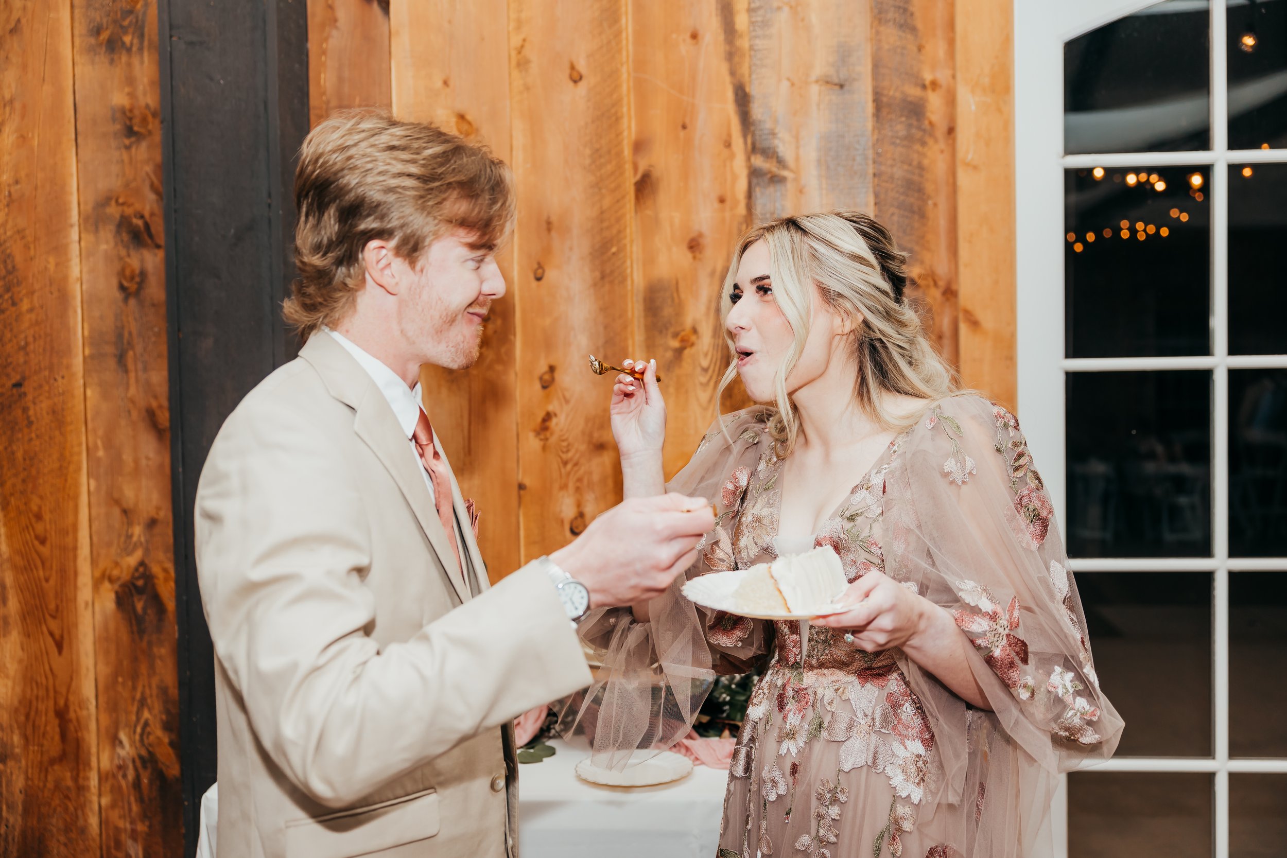 A man and woman sharing a piece of cake at an event in a rustic setting, with a wooden wall and large window in the background.