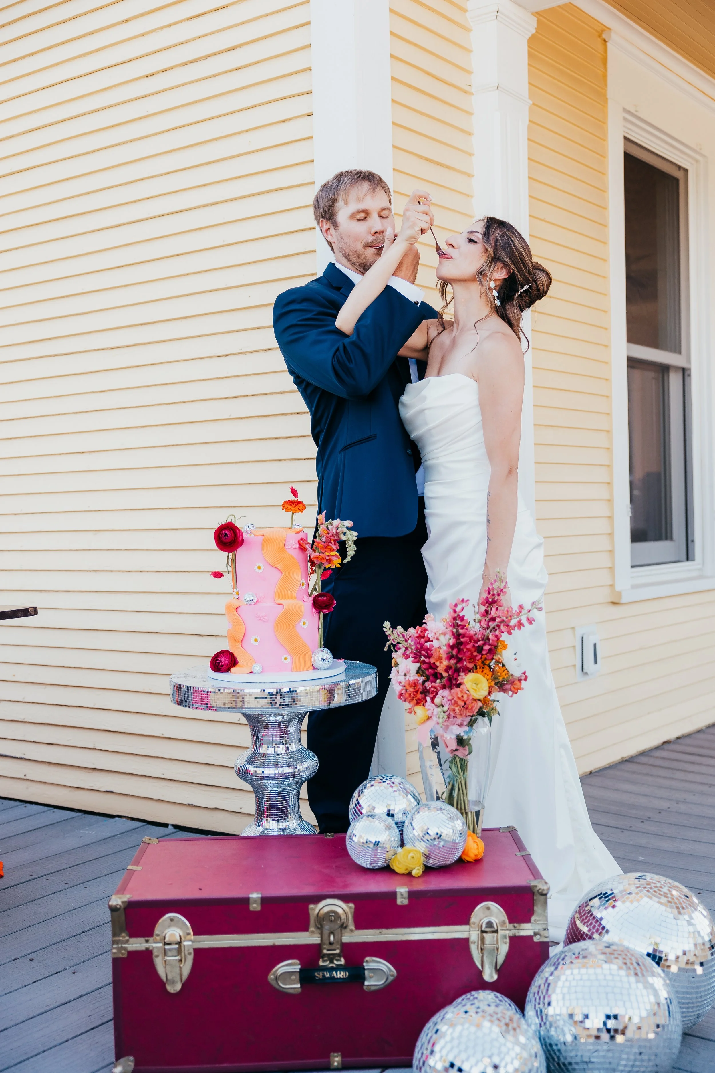 A bride and groom celebrating their wedding outdoors, with the groom feeding the bride cake. There is a pink wedding cake decorated with flowers on a silver stand, a large red trunk, and several disco balls and a bouquet of pink and orange flowers ne
