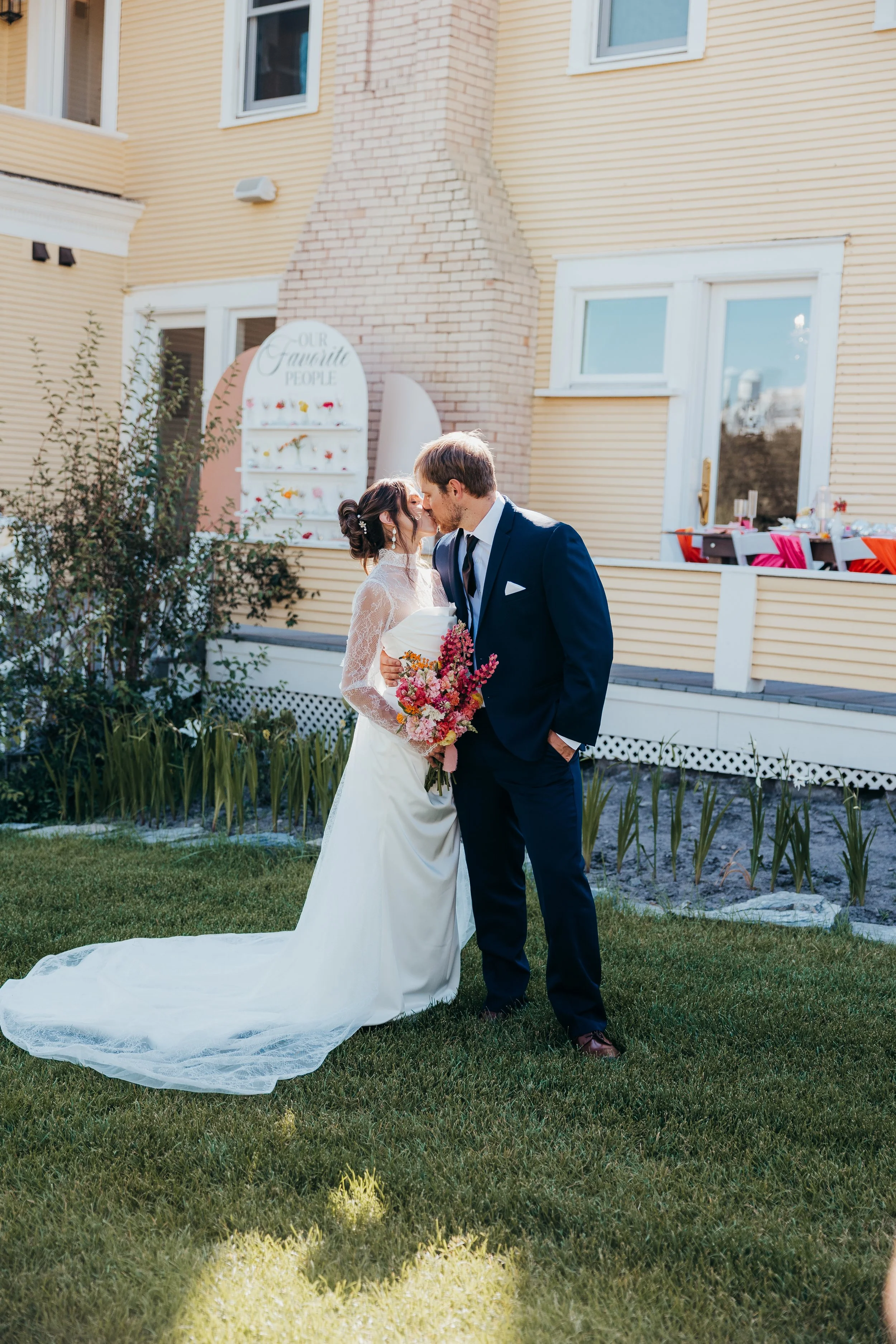 A bride and groom sharing a kiss outdoors during their wedding, with the bride holding a bouquet of flowers. The bride is wearing a white wedding dress with lace sleeves and a long train, while the groom is in a navy suit with a tie. Behind them is a