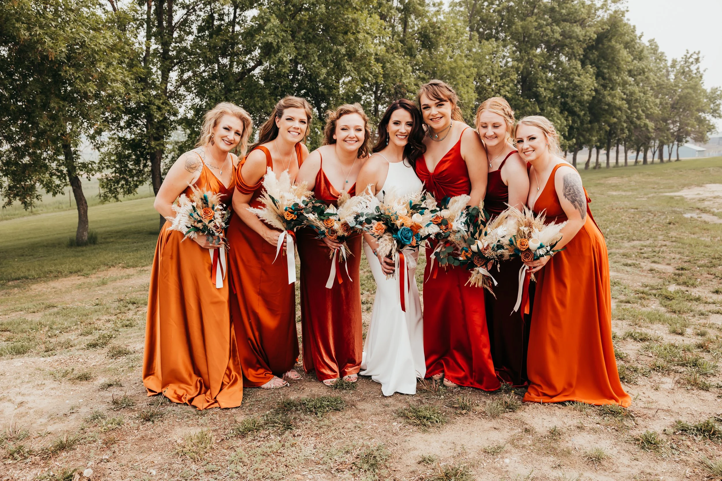 A group of seven women in colorful dresses standing outdoors on a grassy area with trees in the background, each holding a bouquet of flowers.