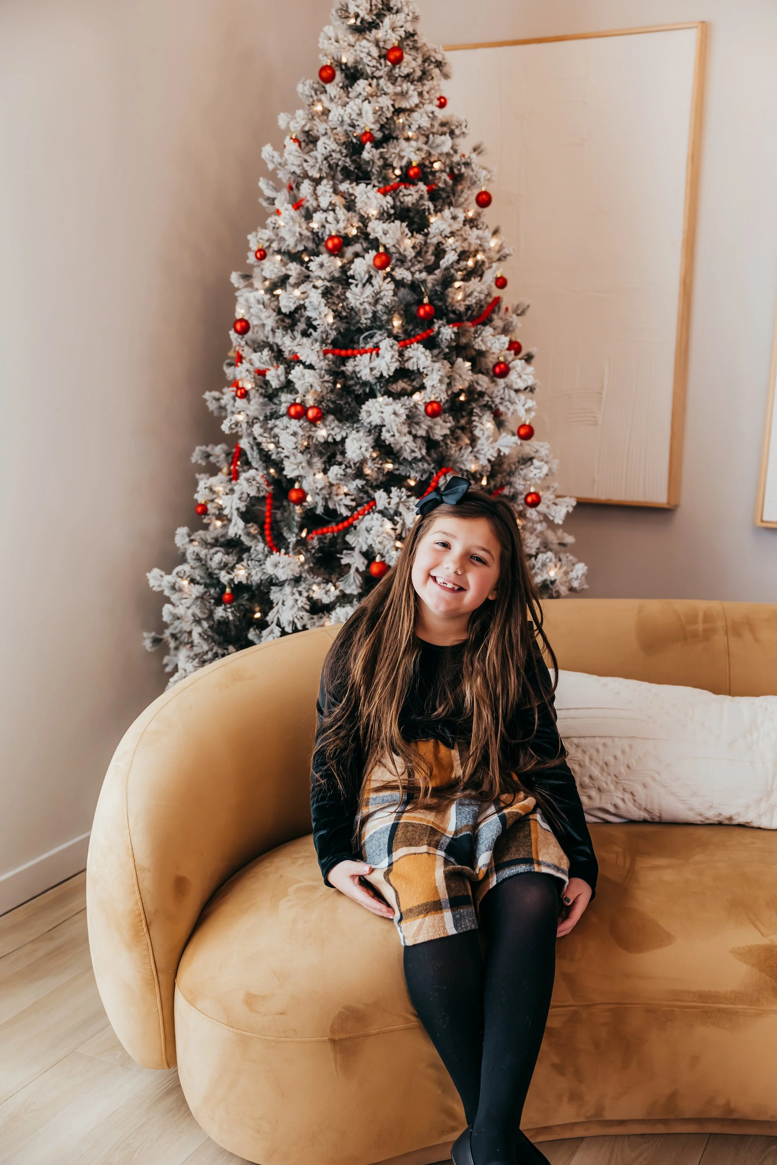 A smiling girl with long brown hair and a black bow sits on a beige sofa in front of a decorated Christmas tree with red ornaments and white flocking.