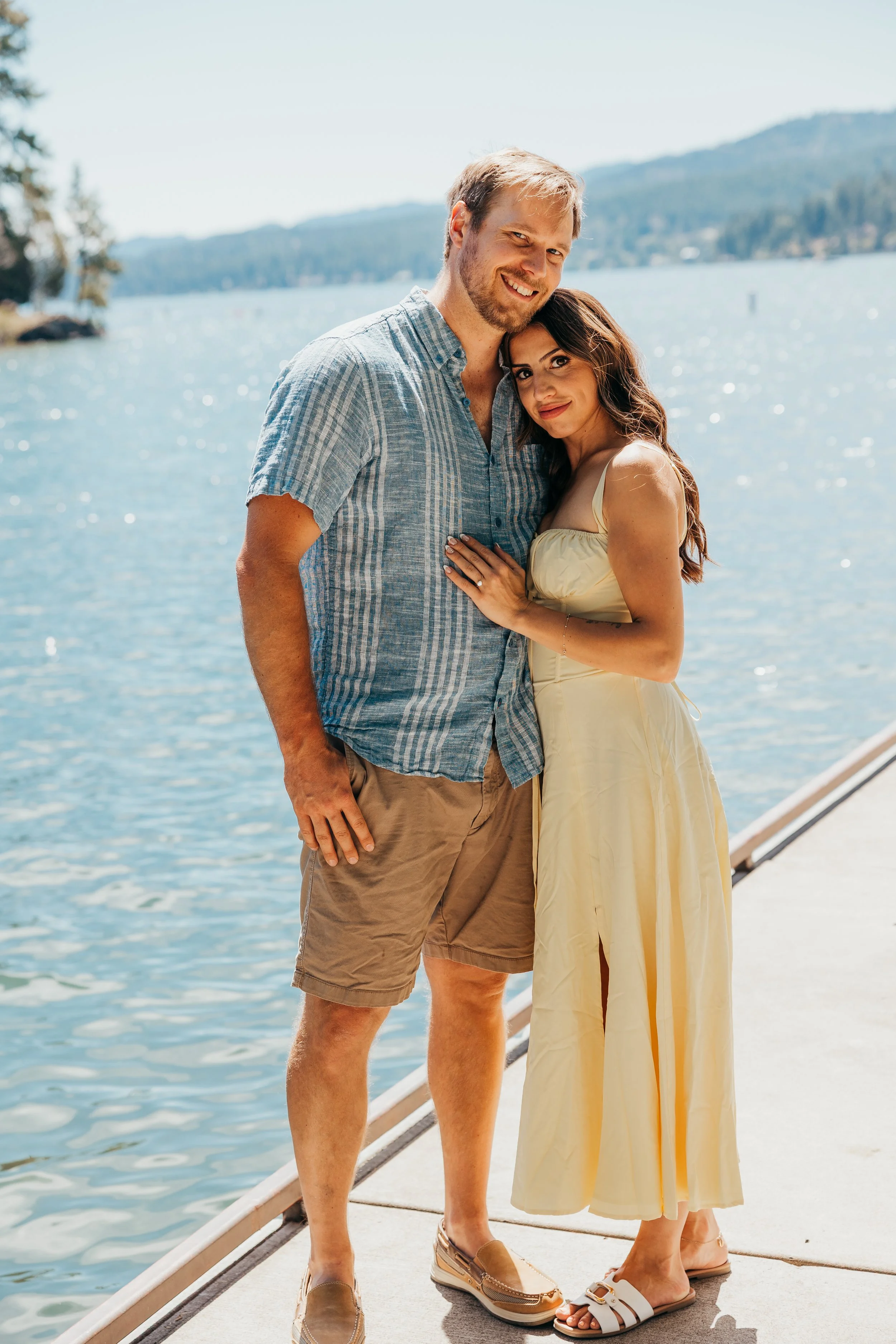 A young couple standing close together on a dock by a lake, smiling and enjoying a sunny day.