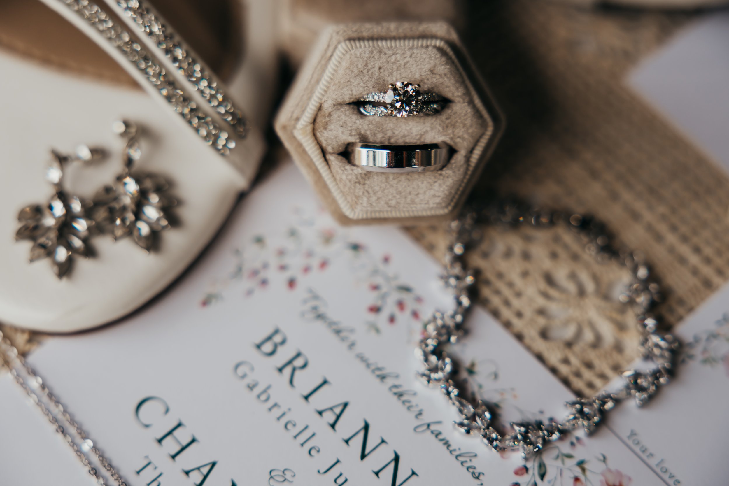 Close-up of a jewelry box with a diamond ring inside, a matching ring below, and nearby jewelry including earrings and a bracelet, with a wedding invitation beneath.