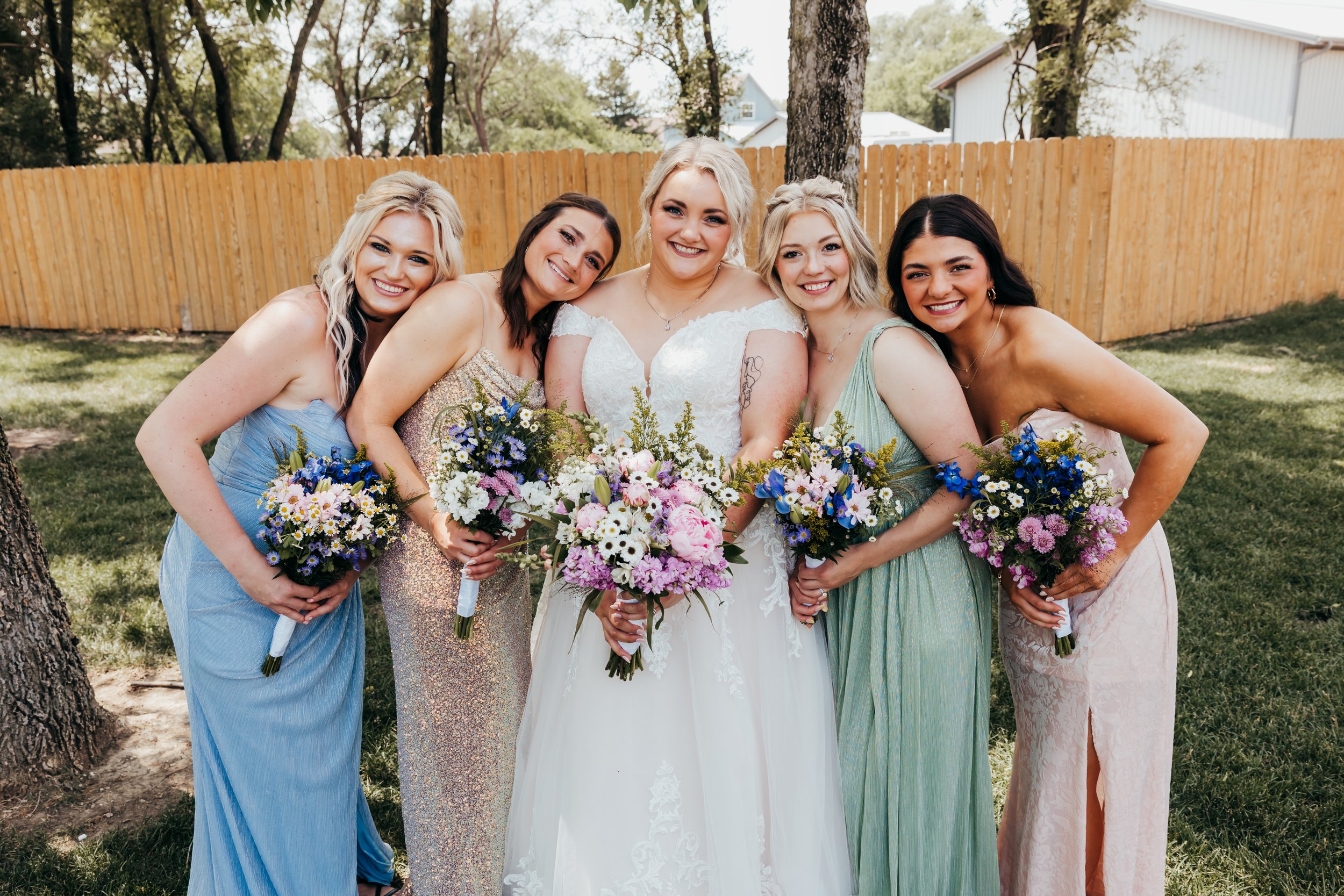 Group of five women standing outdoors, dressed in colorful dresses, holding bouquets of flowers, smiling, with a wooden fence and trees in the background.