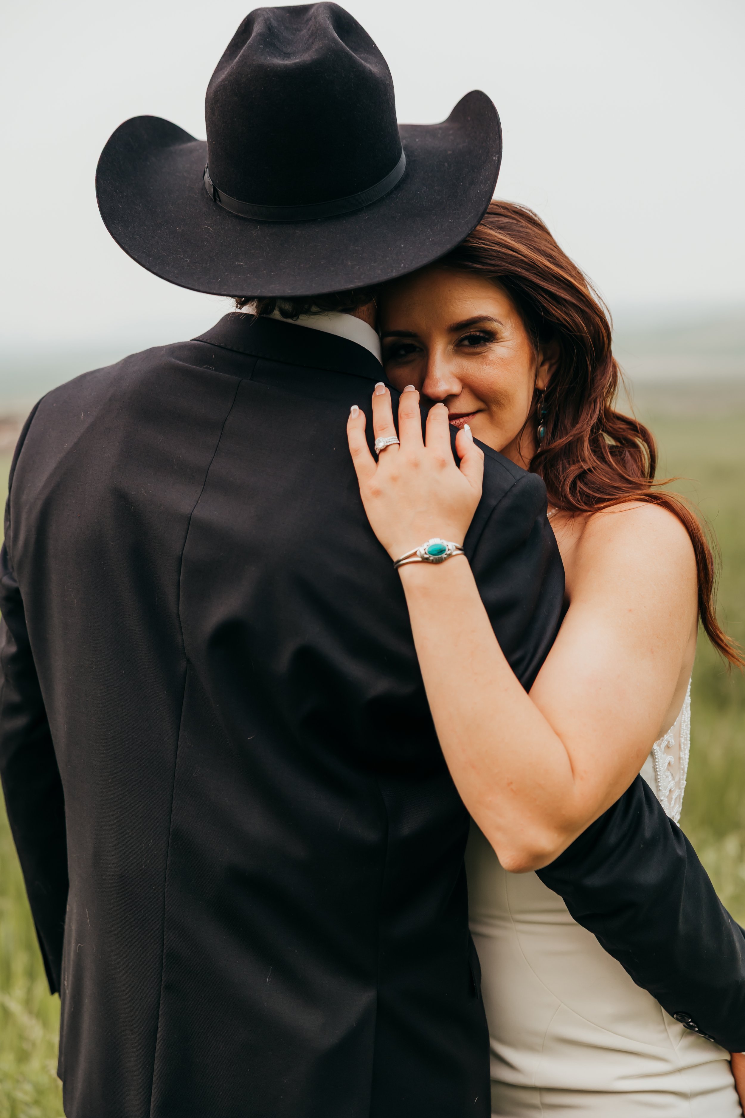 A woman with red hair in a wedding dress hugging a man in a black jacket and black cowboy hat outdoors.