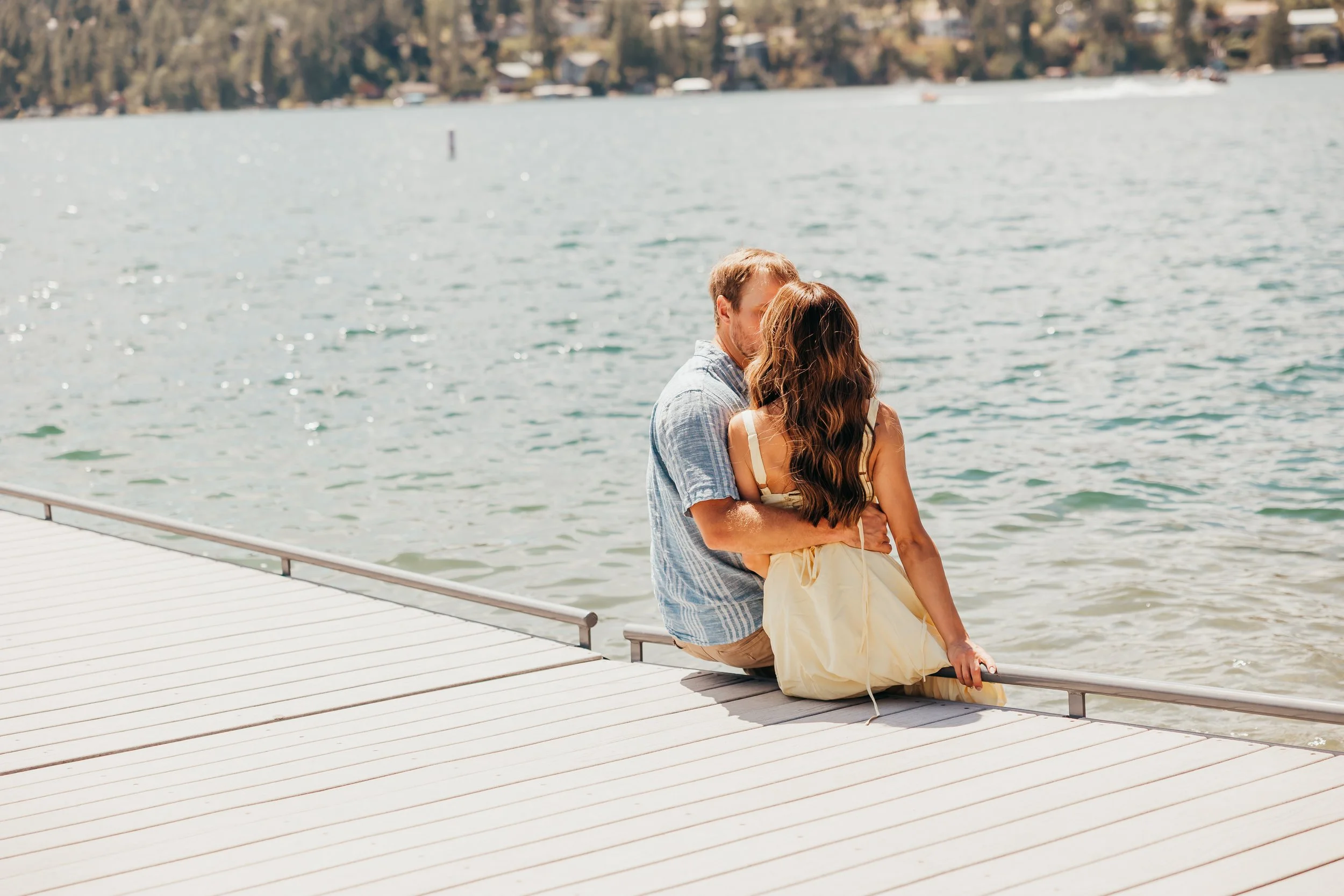 A couple sitting on a dock at the edge of a lake, with the man embracing the woman as they look at the water.