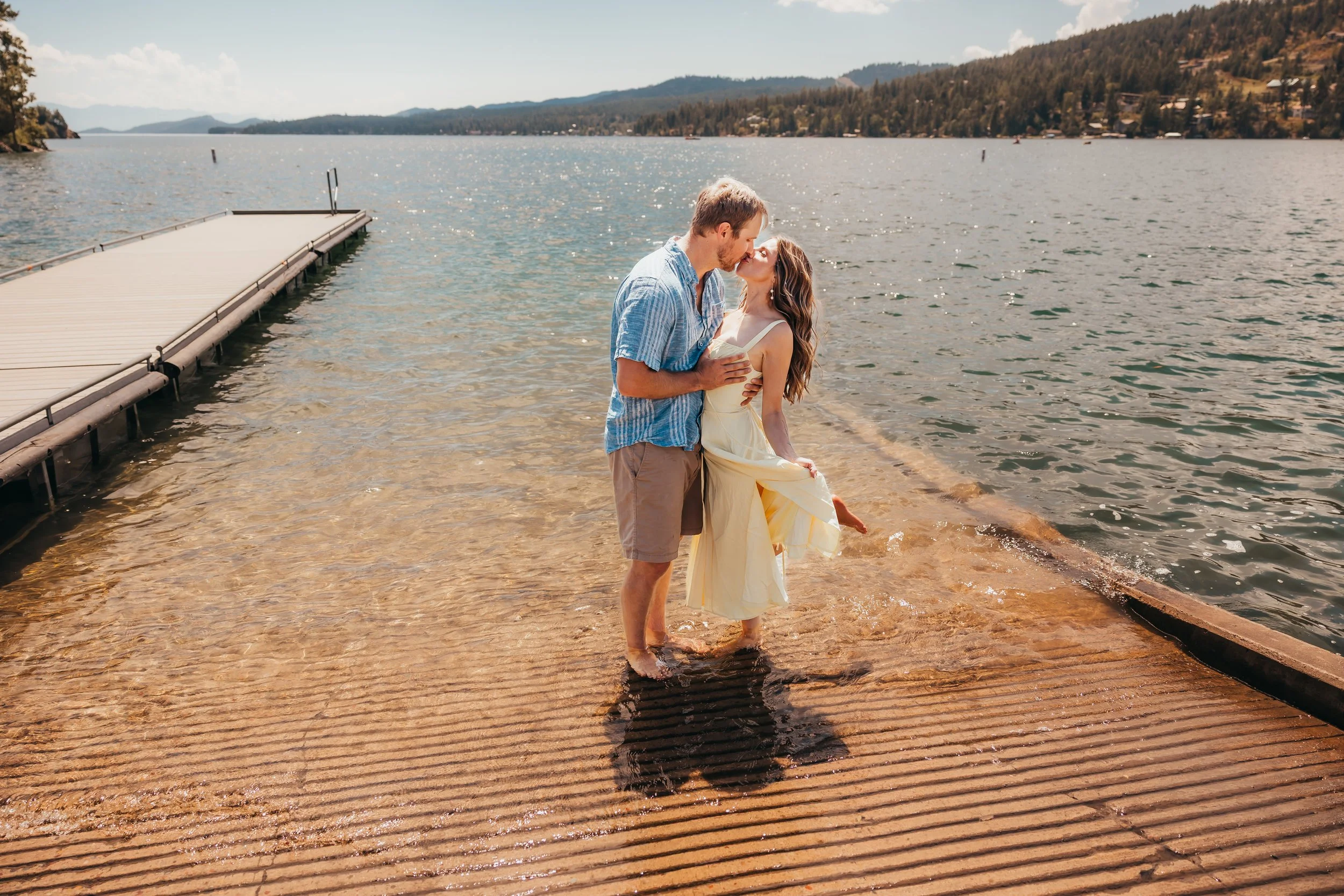 A couple standing on a wooden dock by a lake, sharing a kiss. The man is wearing a blue shirt and shorts, and the woman is wearing a yellow dress. It is a sunny day with mountains and trees in the background.