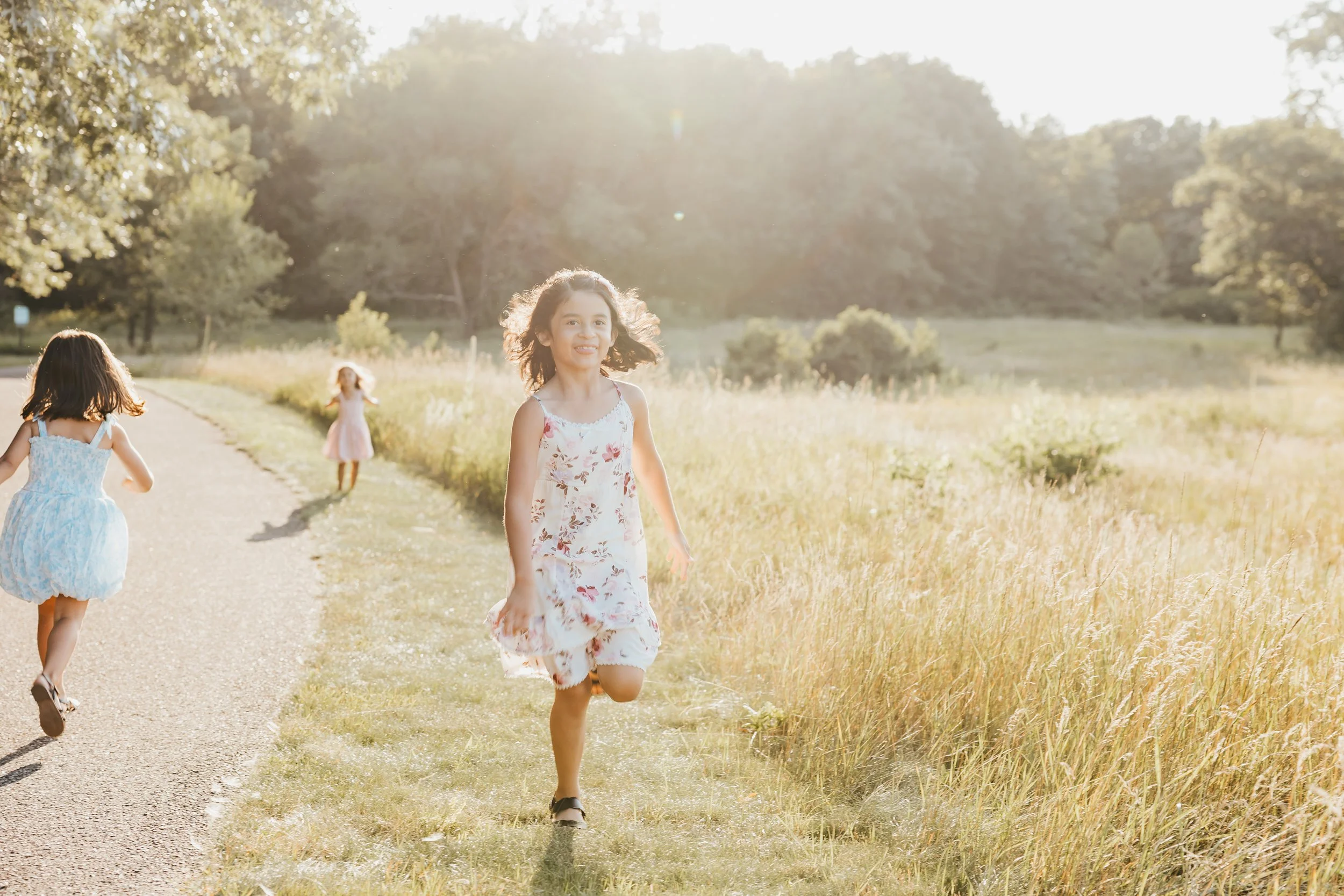 Three young girls in dresses playing and running along a country path during sunset, surrounded by tall grass and trees.