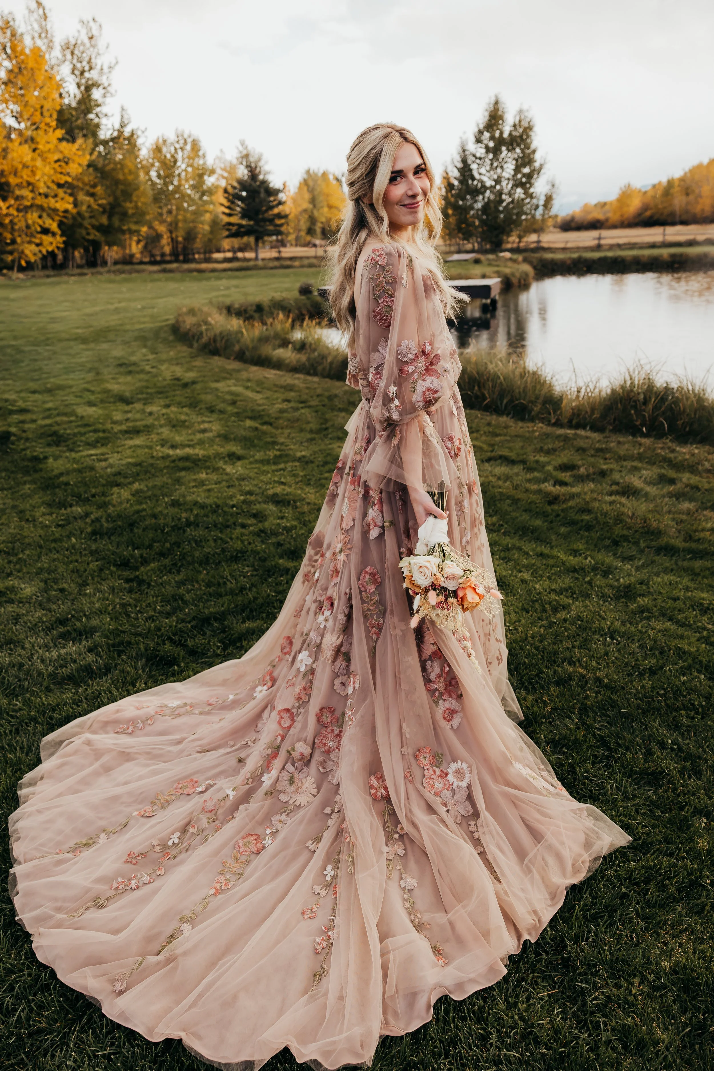 A woman in a long, pink, embroidered dress holding a bouquet of flowers, standing on a grassy bank near a pond with trees in the background during fall.