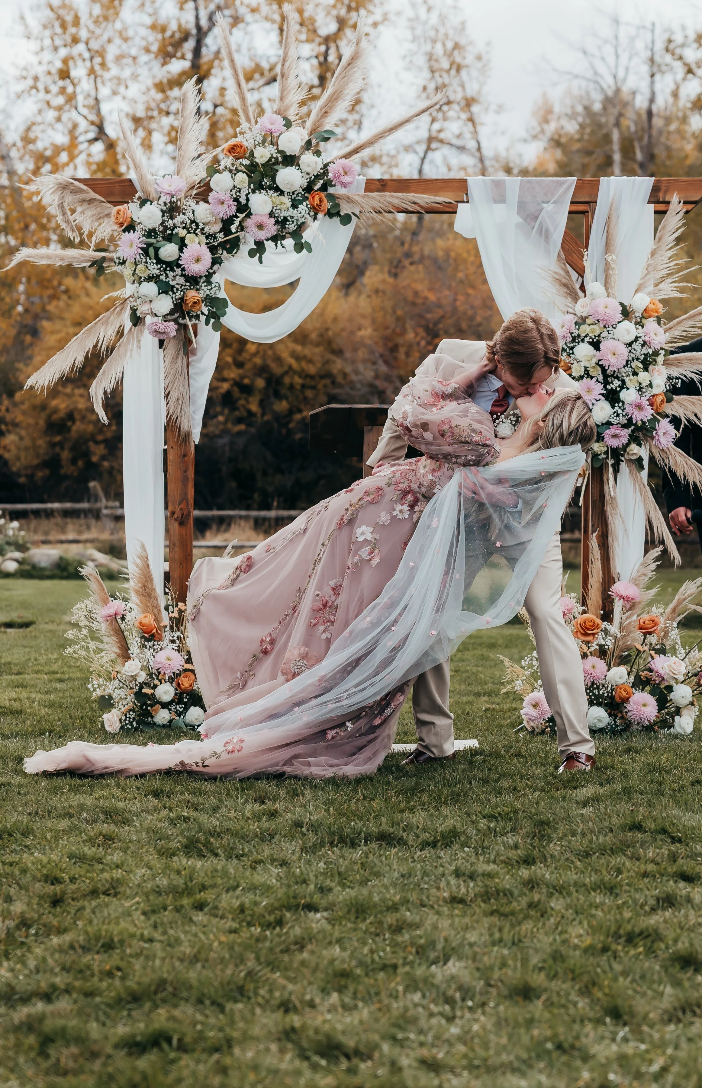 Couple sharing a kiss during their outdoor wedding ceremony with floral decorations and a wooden arch.