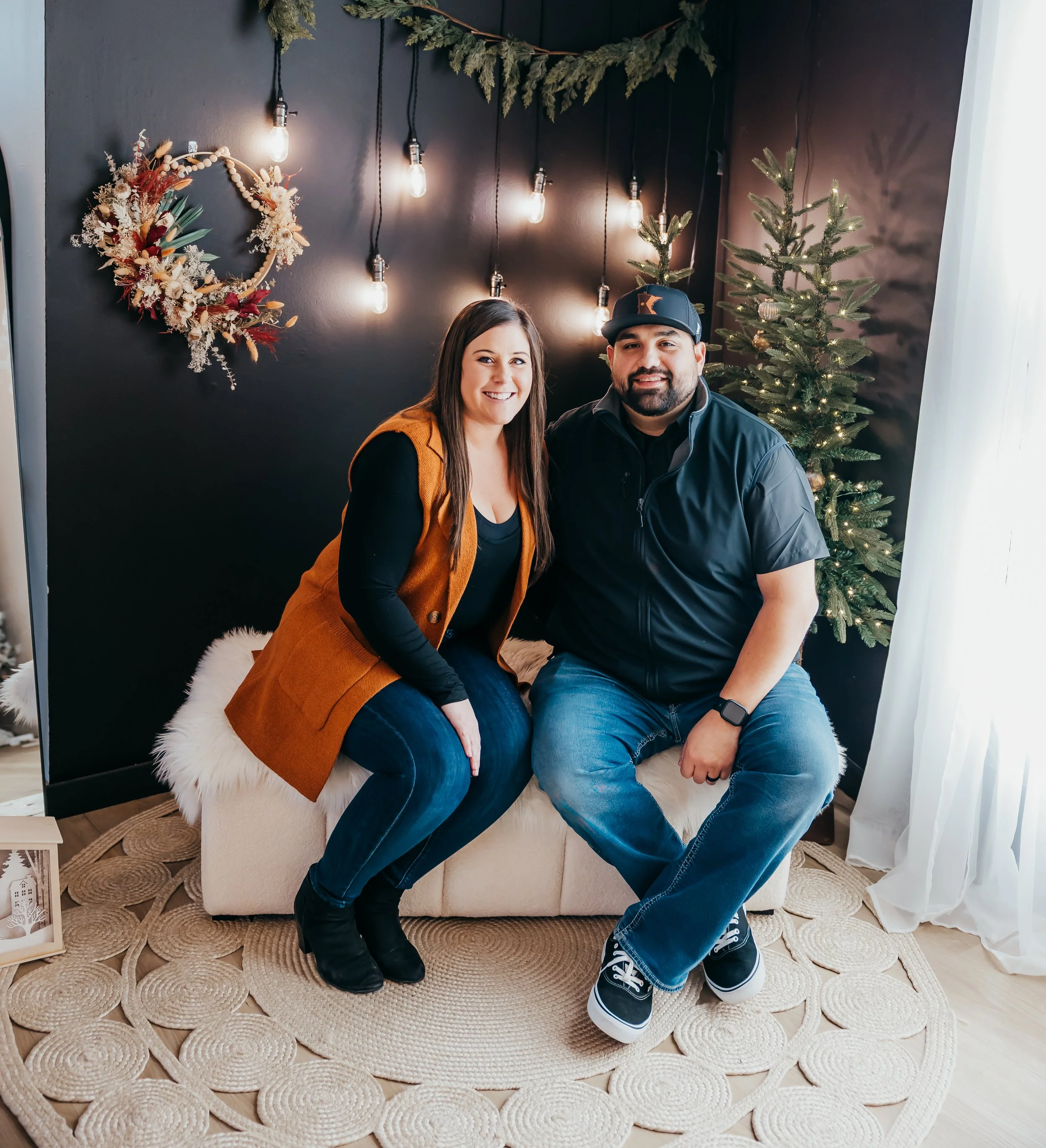 A woman and a man sitting on a cozy bench in a festively decorated room with Christmas decorations, including a small Christmas tree, a decorative wreath, and string lights.
