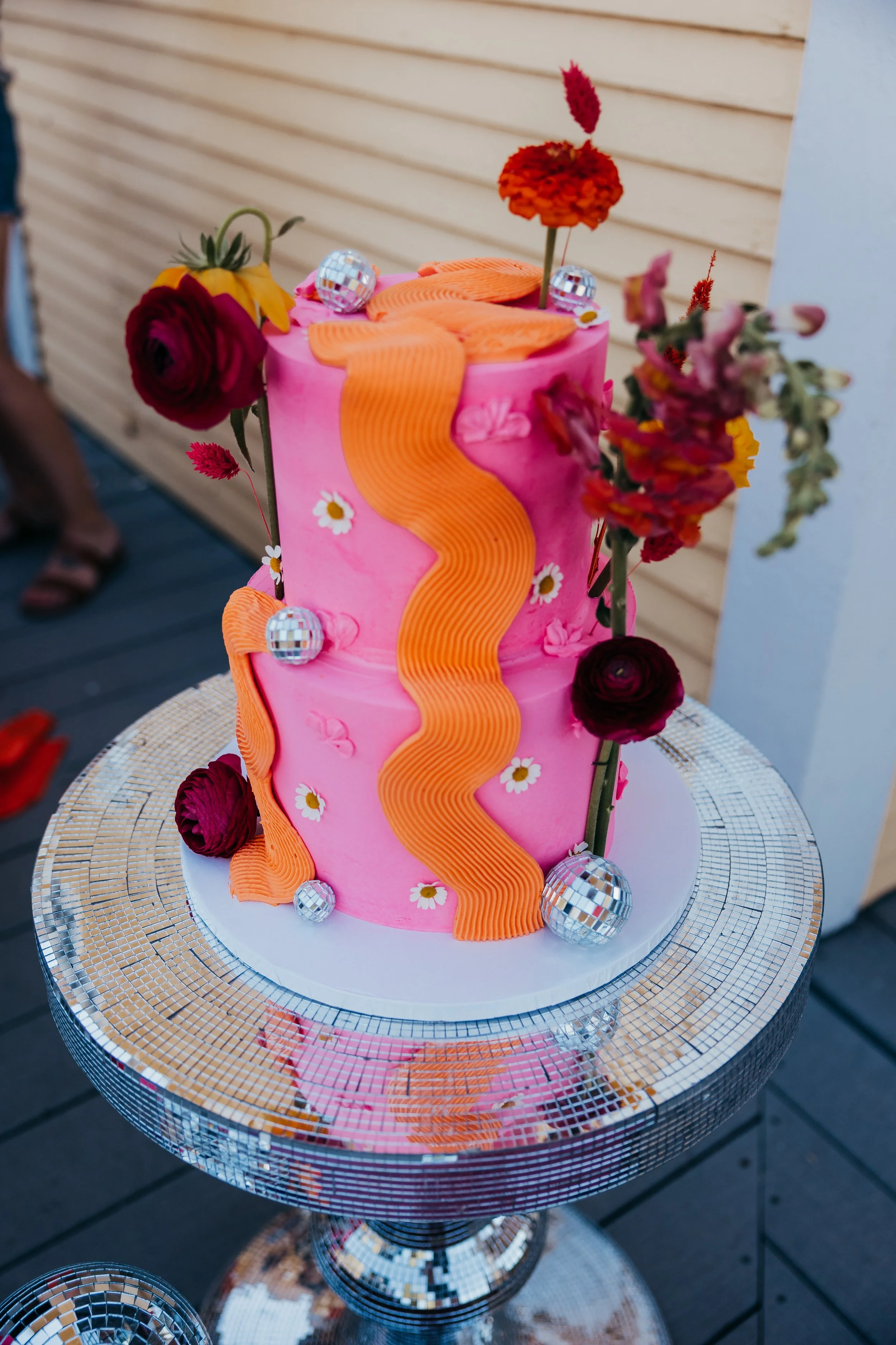 A pink two-tiered cake decorated with orange streamers, real flowers, disco balls, and small daisy flowers, displayed on a reflective silver stand.