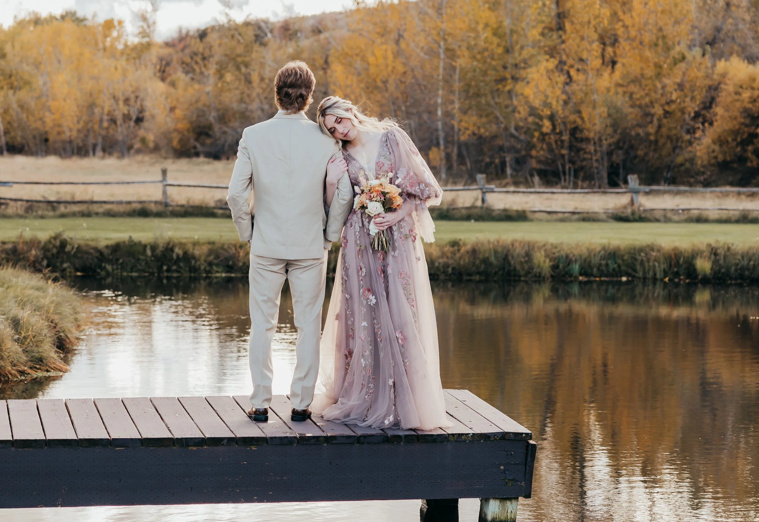 A man in a light-colored suit and a woman in a floral pink and lavender gown standing on a wooden dock by a lake, holding hands, with a fall landscape of trees with orange and yellow leaves in the background.