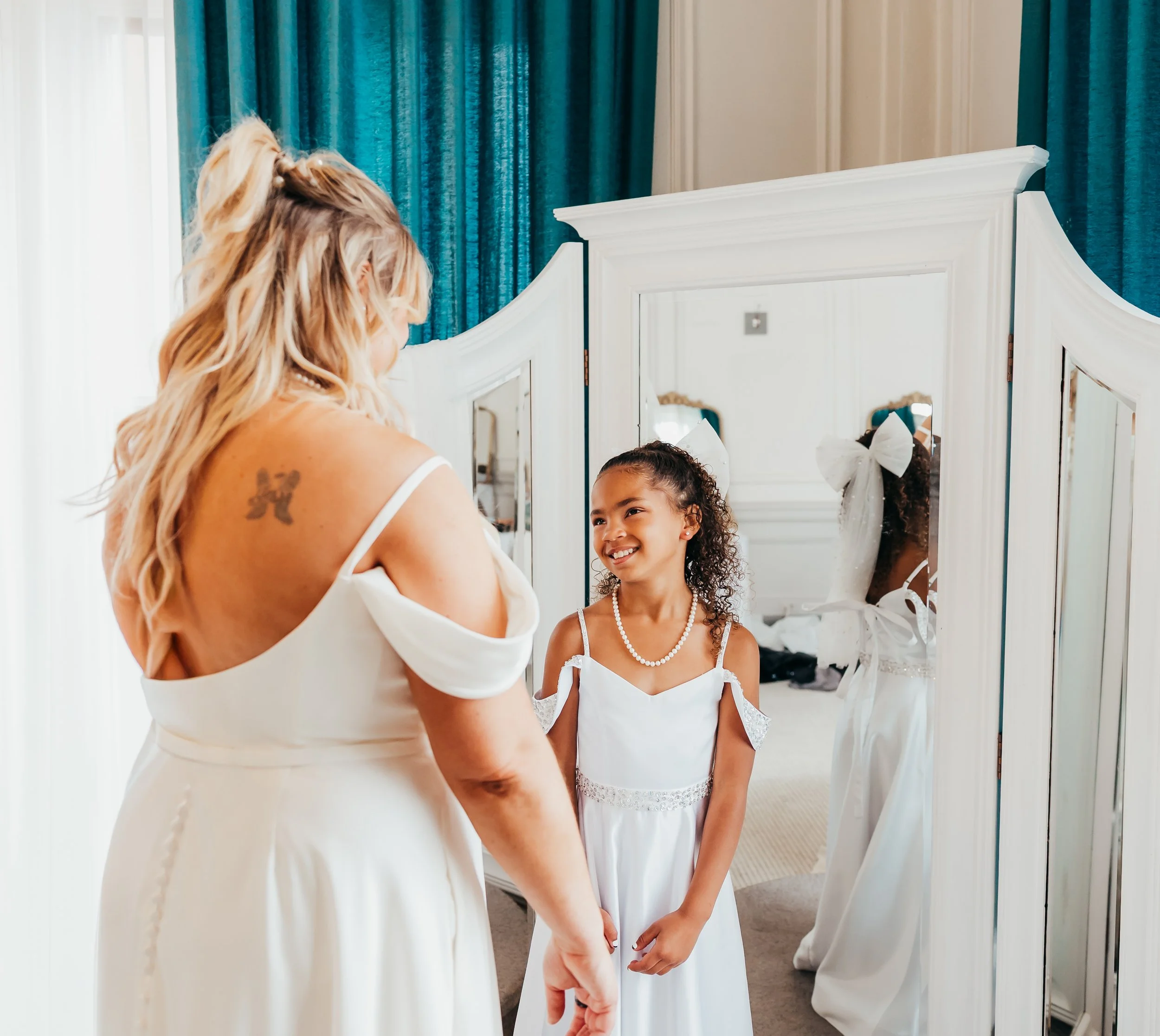 A woman and a girl smiling at each other in front of a mirror, both dressed in white gowns, likely getting ready for a special occasion in a room with blue curtains.