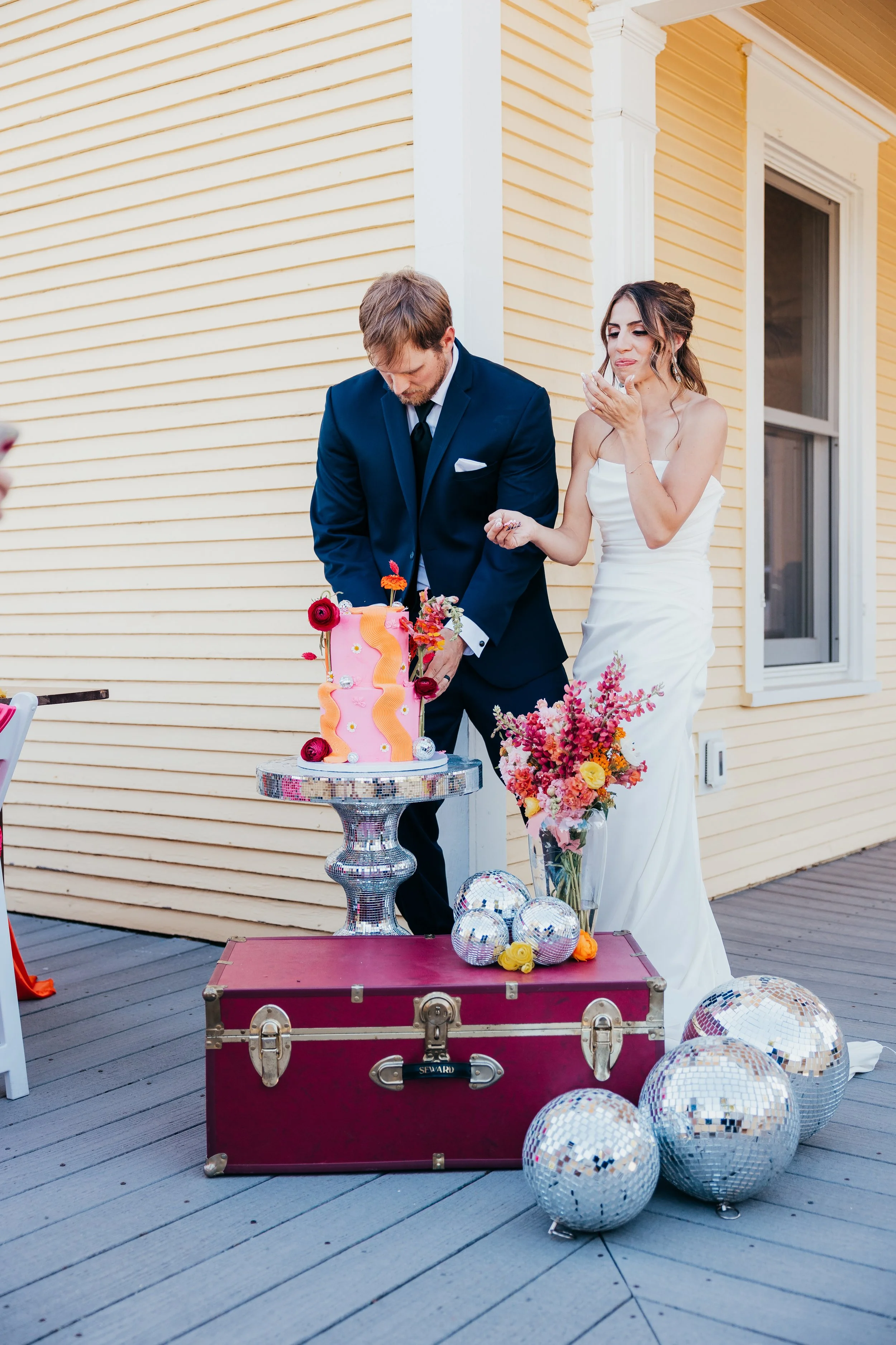 A bride and groom at a wedding celebration cutting a colorful wedding cake outdoors near a yellow house. The bride is in a white strapless gown, and the groom is in a dark suit with a tie. There are disco balls and a large pink floral gift box on the