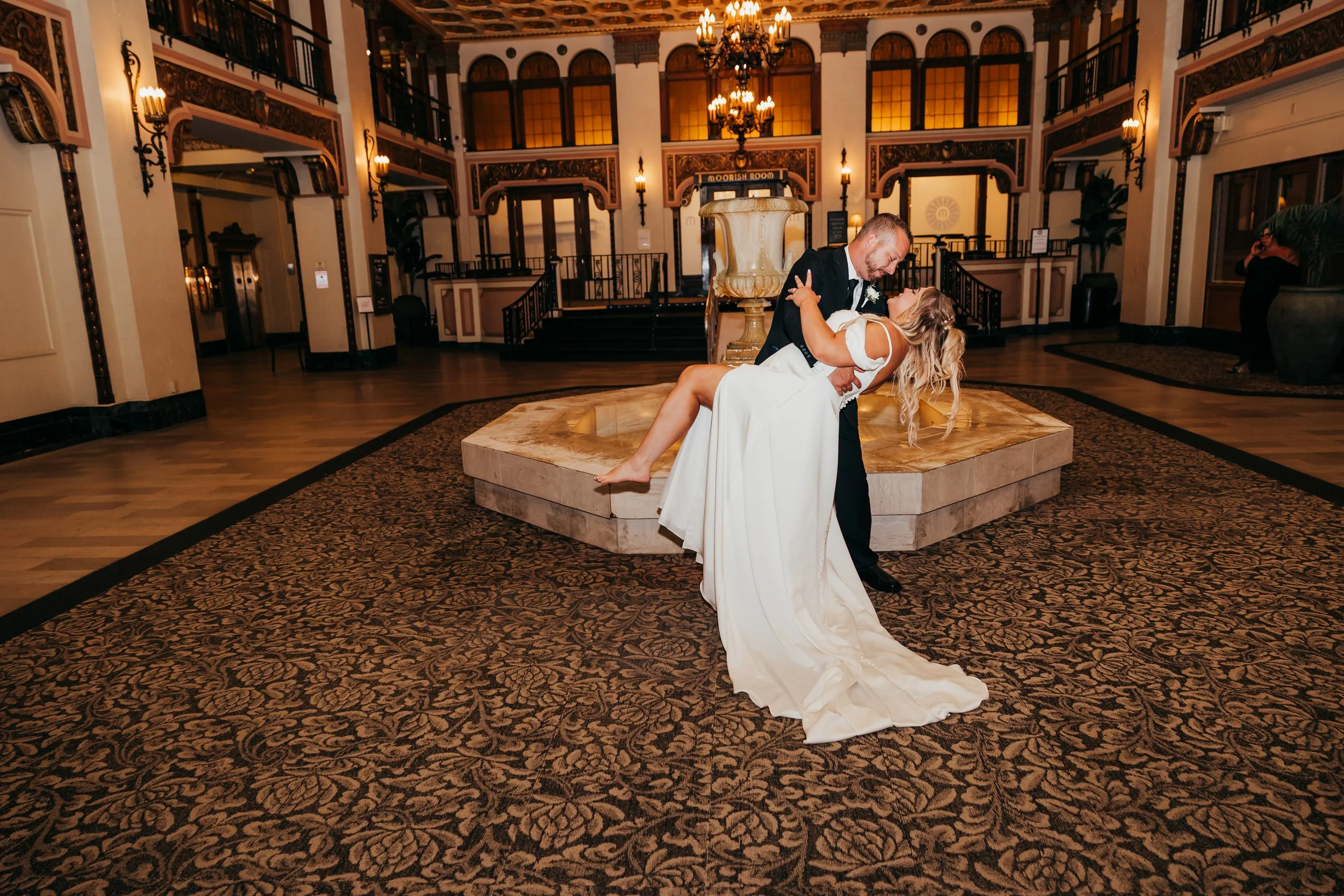 Bride and groom dancing in a grand, ornate hall with warm lighting and elegant decor.