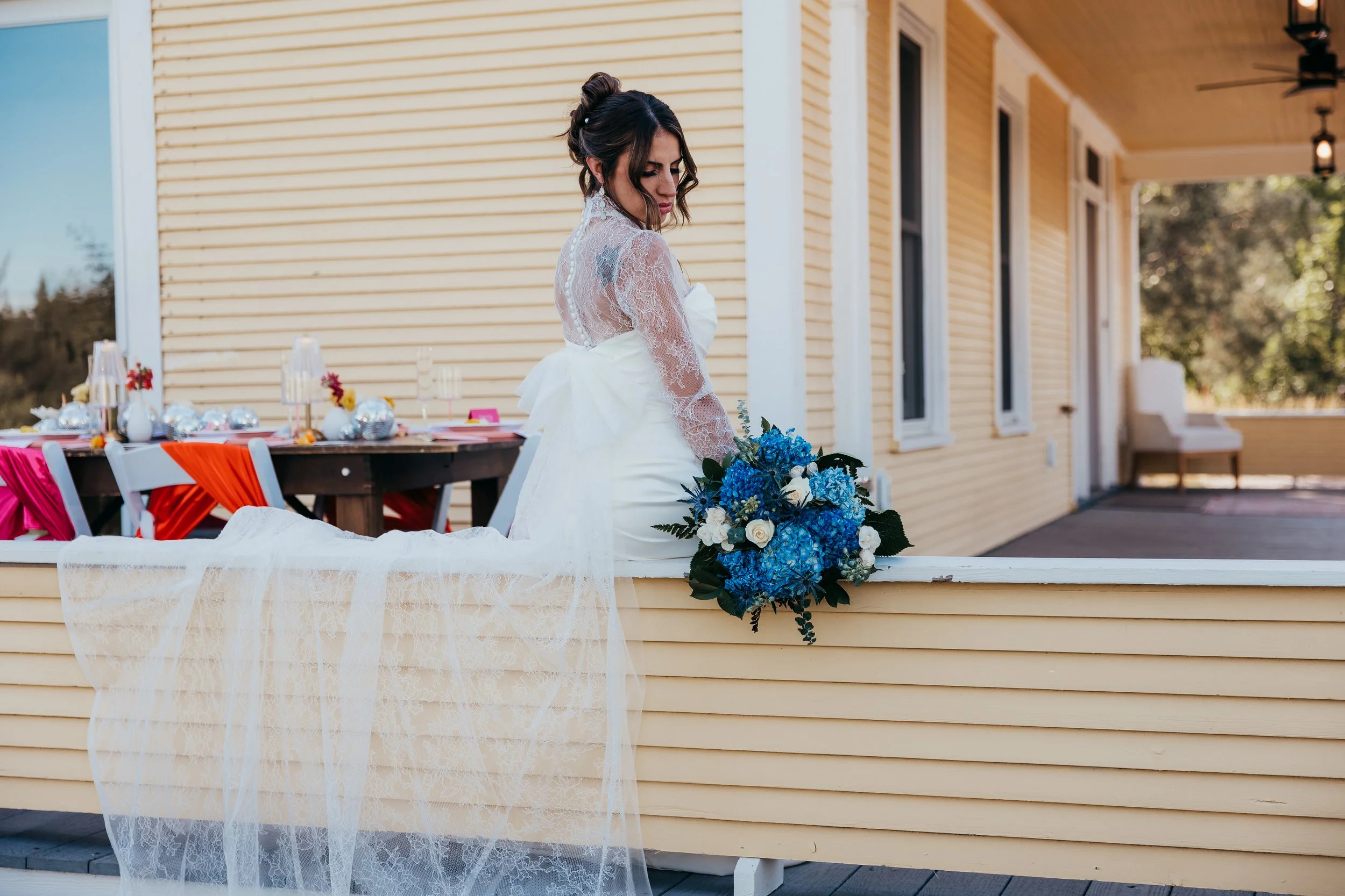 Bride in a white wedding dress with lace sleeves and veil holding a blue and white flower bouquet on a porch with yellow siding.