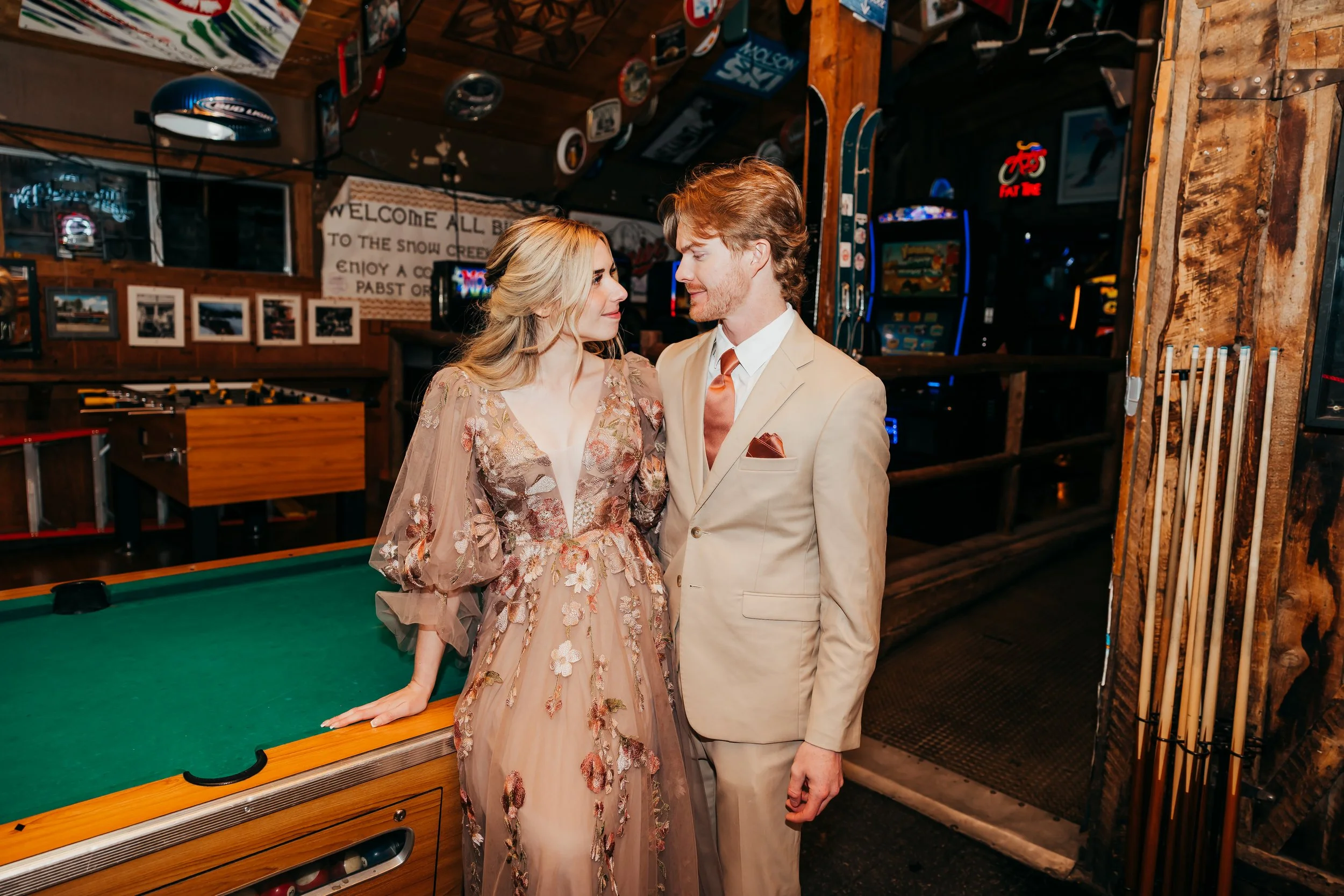 A man and woman in formal attire inside a rustic bar, looking at each other, with pool tables and arcade games in the background.