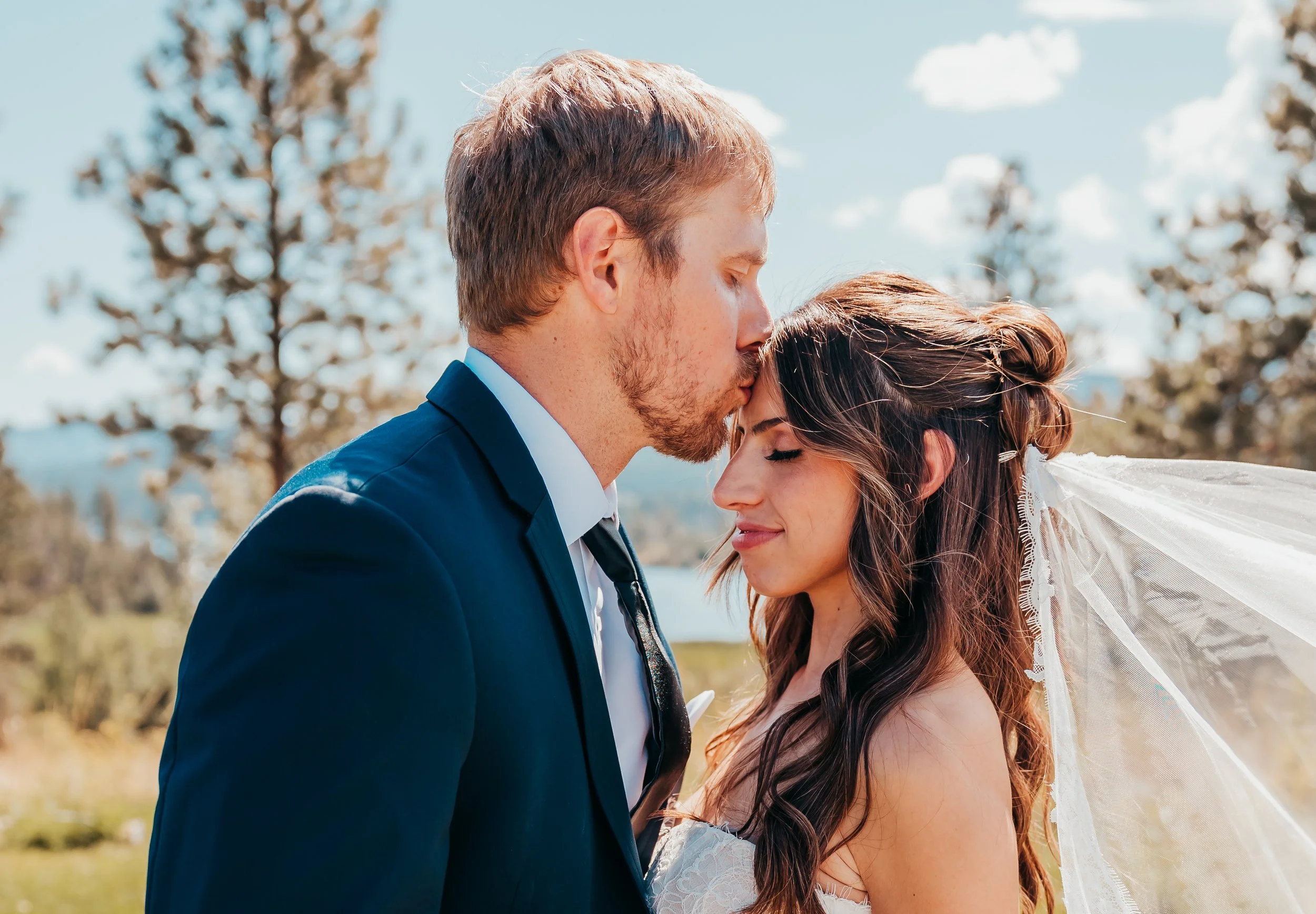 A bride and groom share an intimate moment outdoors on their wedding day, with the groom kissing the bride's forehead and the bride smiling softly with eyes closed.