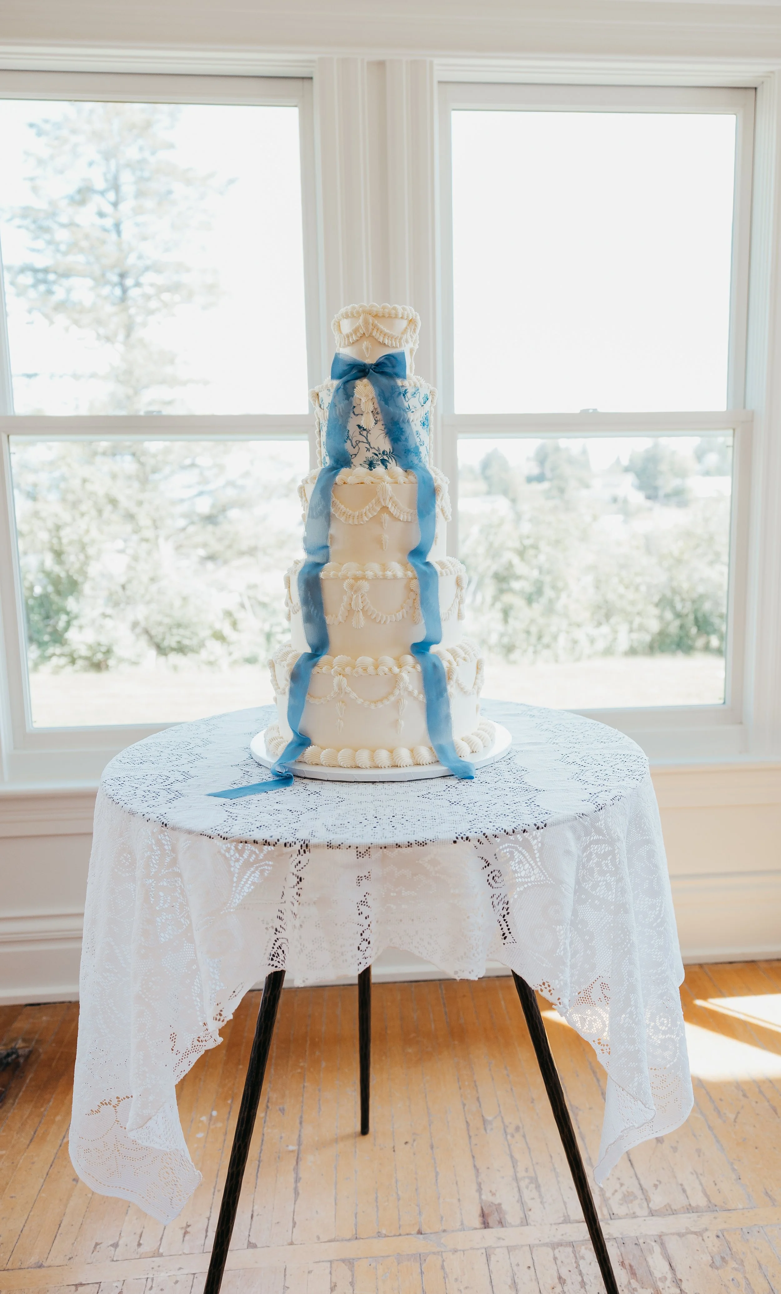 Four-tier white wedding cake with blue ribbon decorations, placed on a round table with lace tablecloth in front of large windows.
