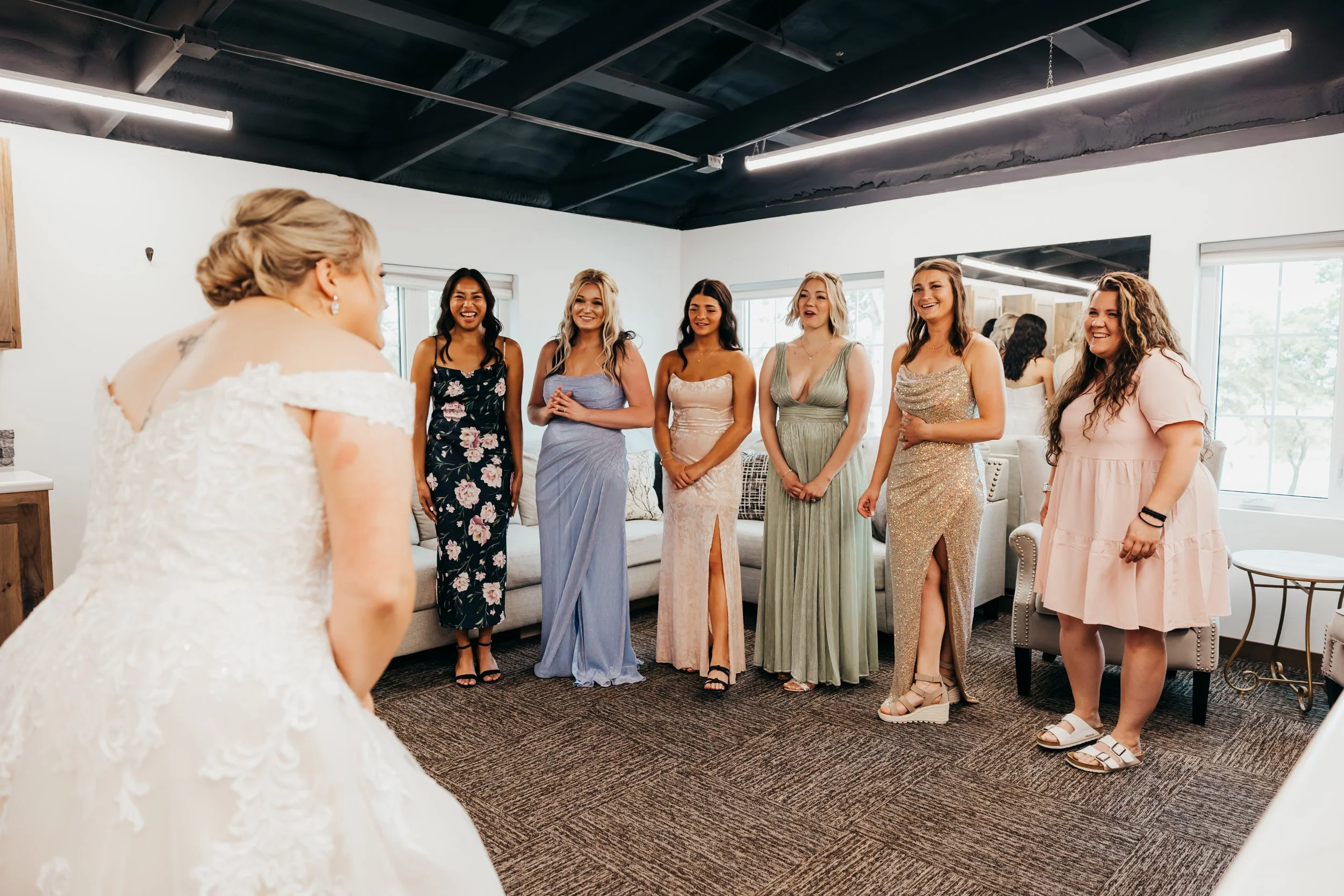 Bride in wedding dress bowing to six women in colorful dresses in a bright room.