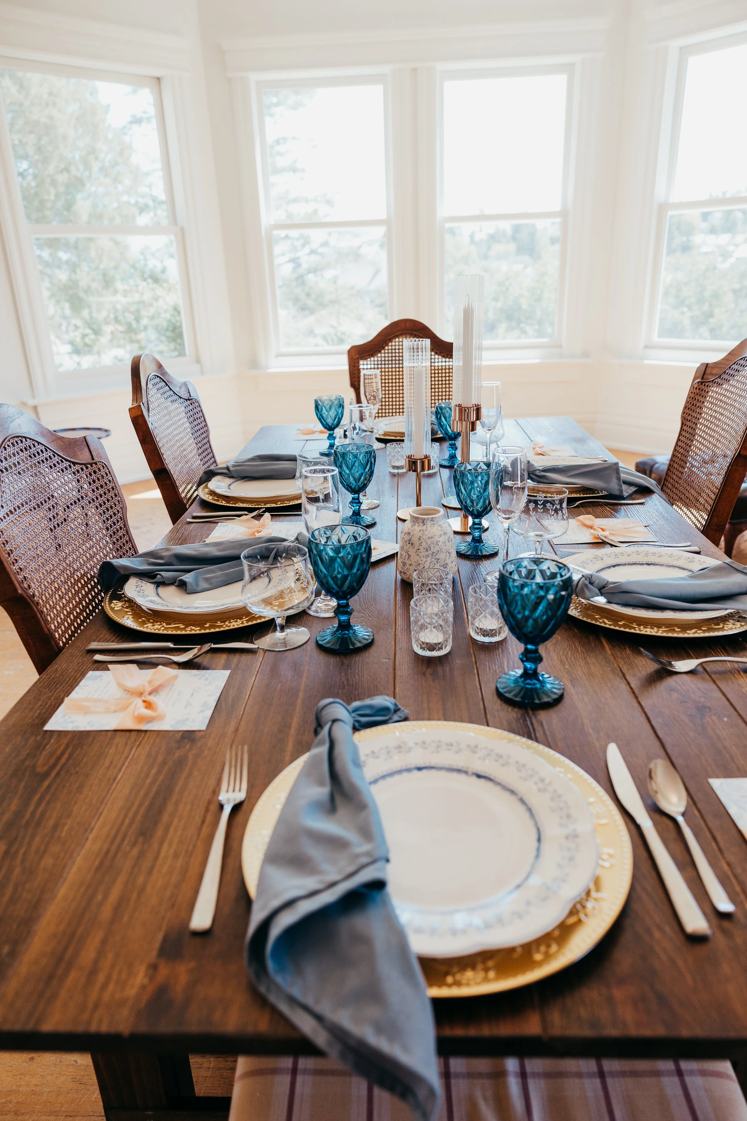 An elegantly set dining table with plates, glasses, blue goblets, napkins, and candles in a bright room.
