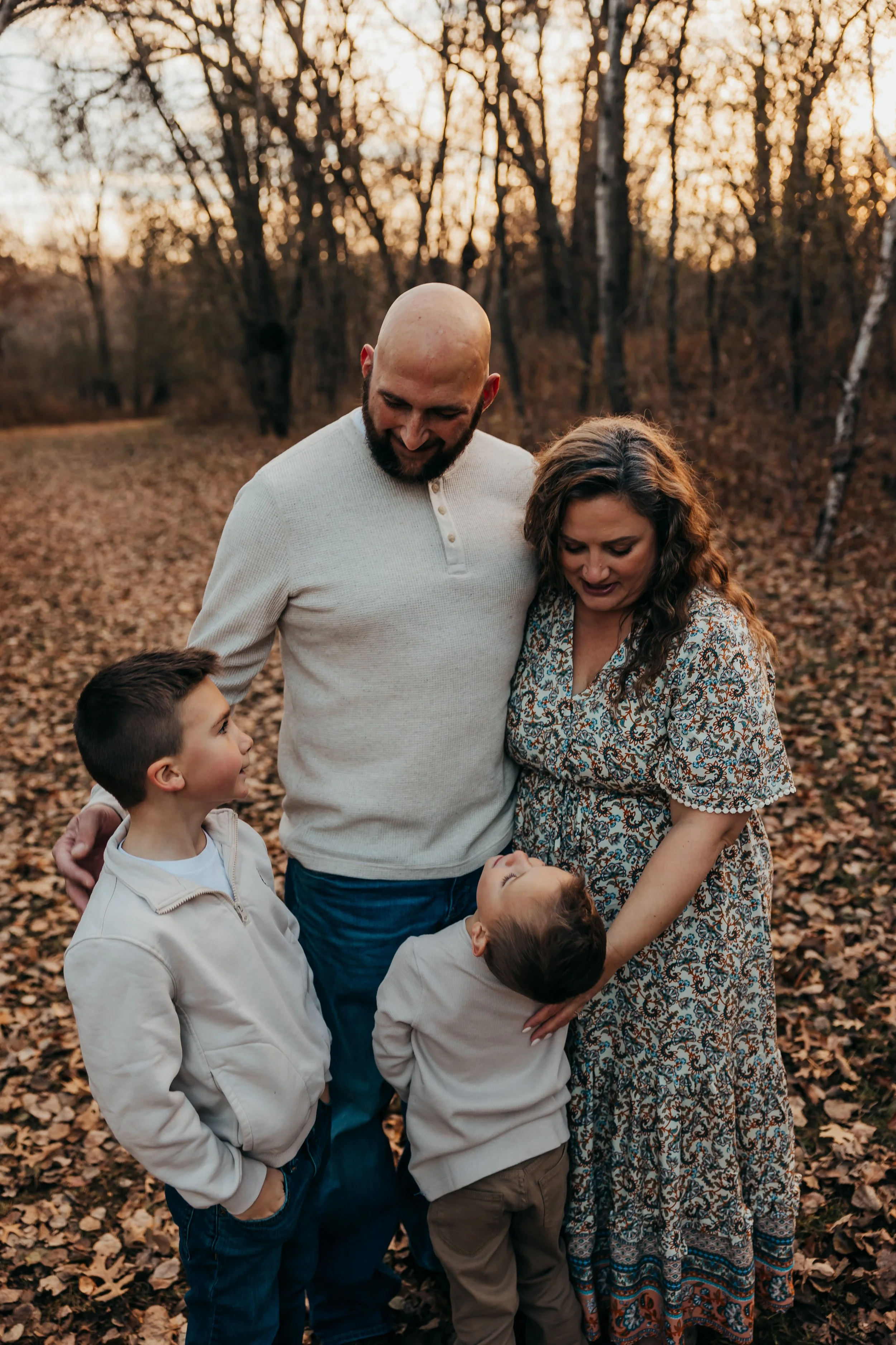 A family of four standing close together in a forest with fallen autumn leaves, smiling and looking at each other as the sun sets behind trees.