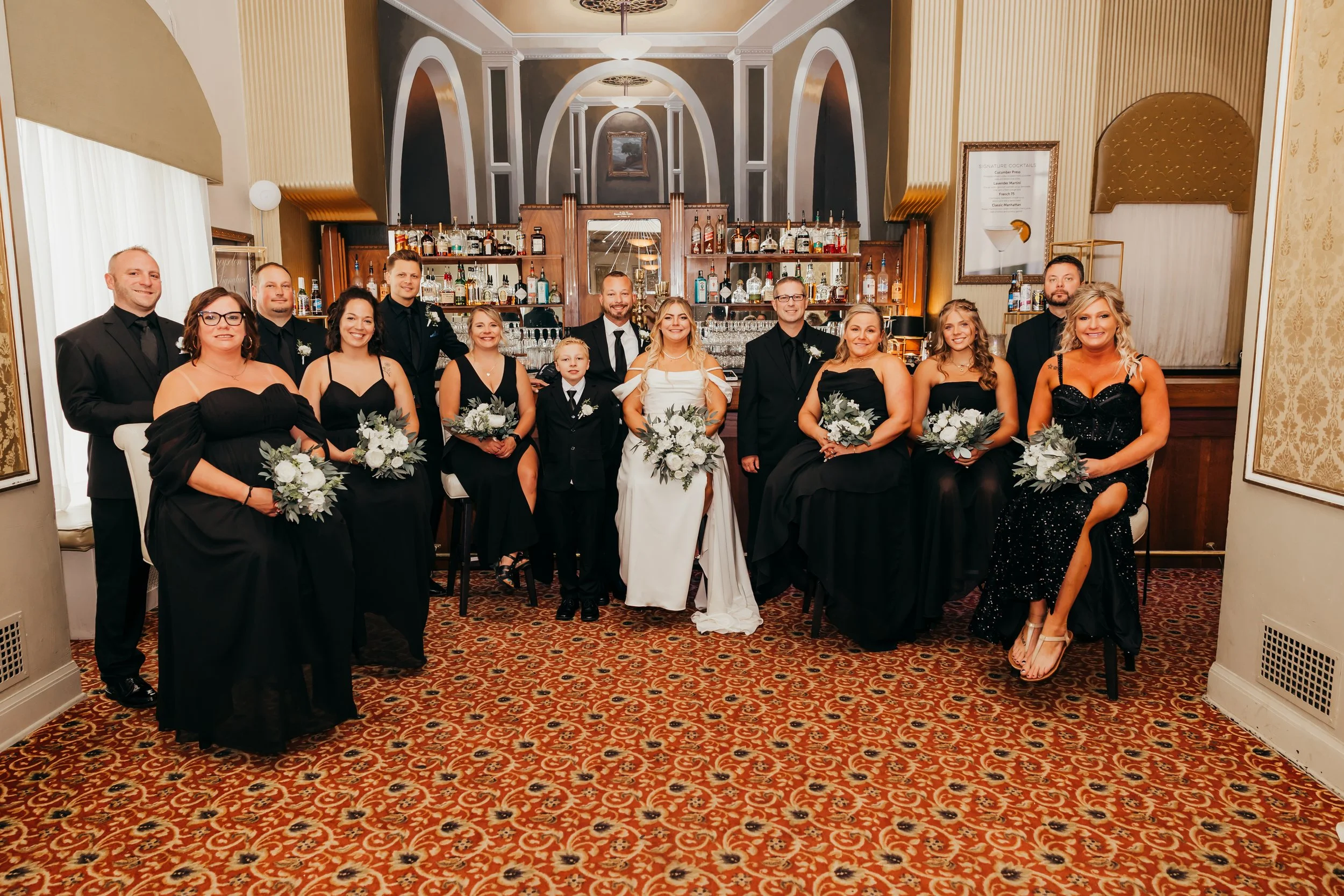 A wedding party with the bride, groom, and bridesmaids and groomsmen posing in a hotel or banquet hall bar area. Women in black dresses hold bouquets, men in black suits. The bride wears a white gown, the groom a black tux. The background features a 