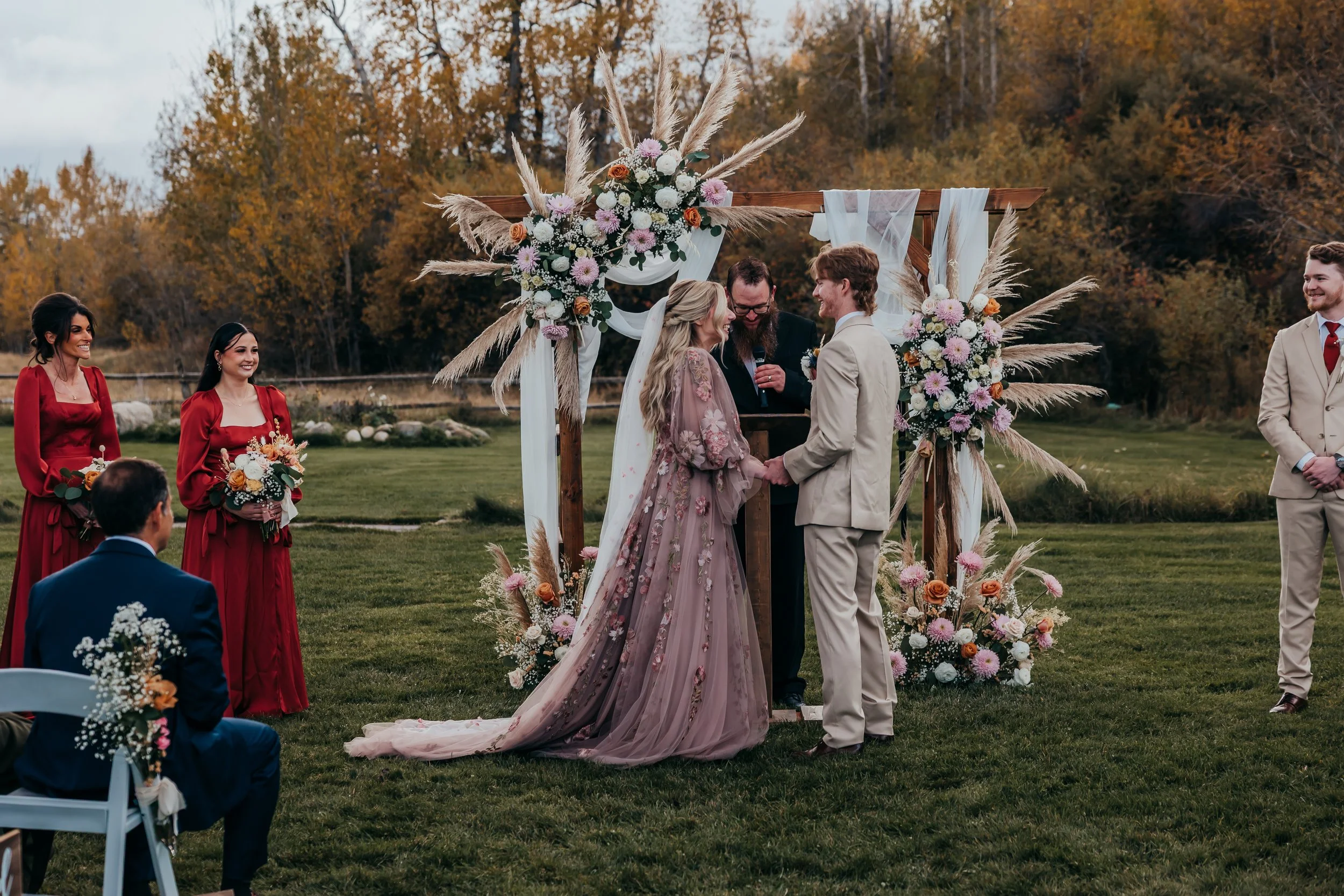 A couple getting married outdoors under a floral arch, with bridesmaids and groomsmen standing nearby, in a grassy area with trees in fall colors.
