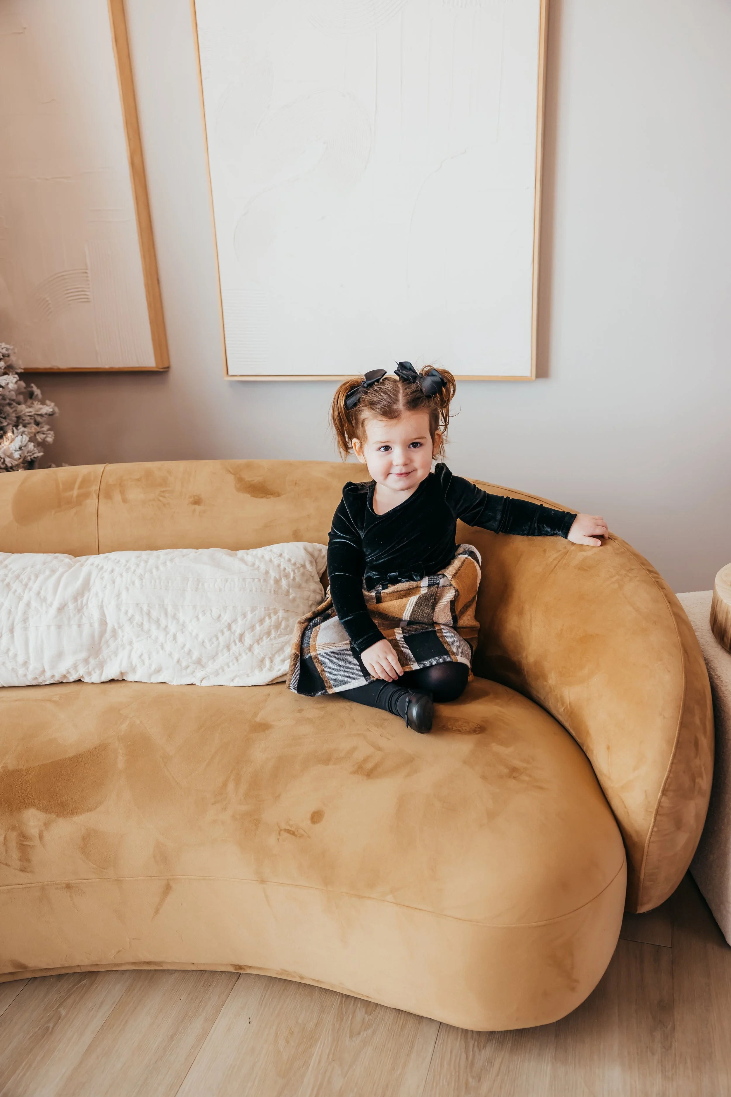 A young girl with brown hair in pigtails and wearing a black dress, plaid skirt, and black tights, sitting on a tan sofa in a living room.