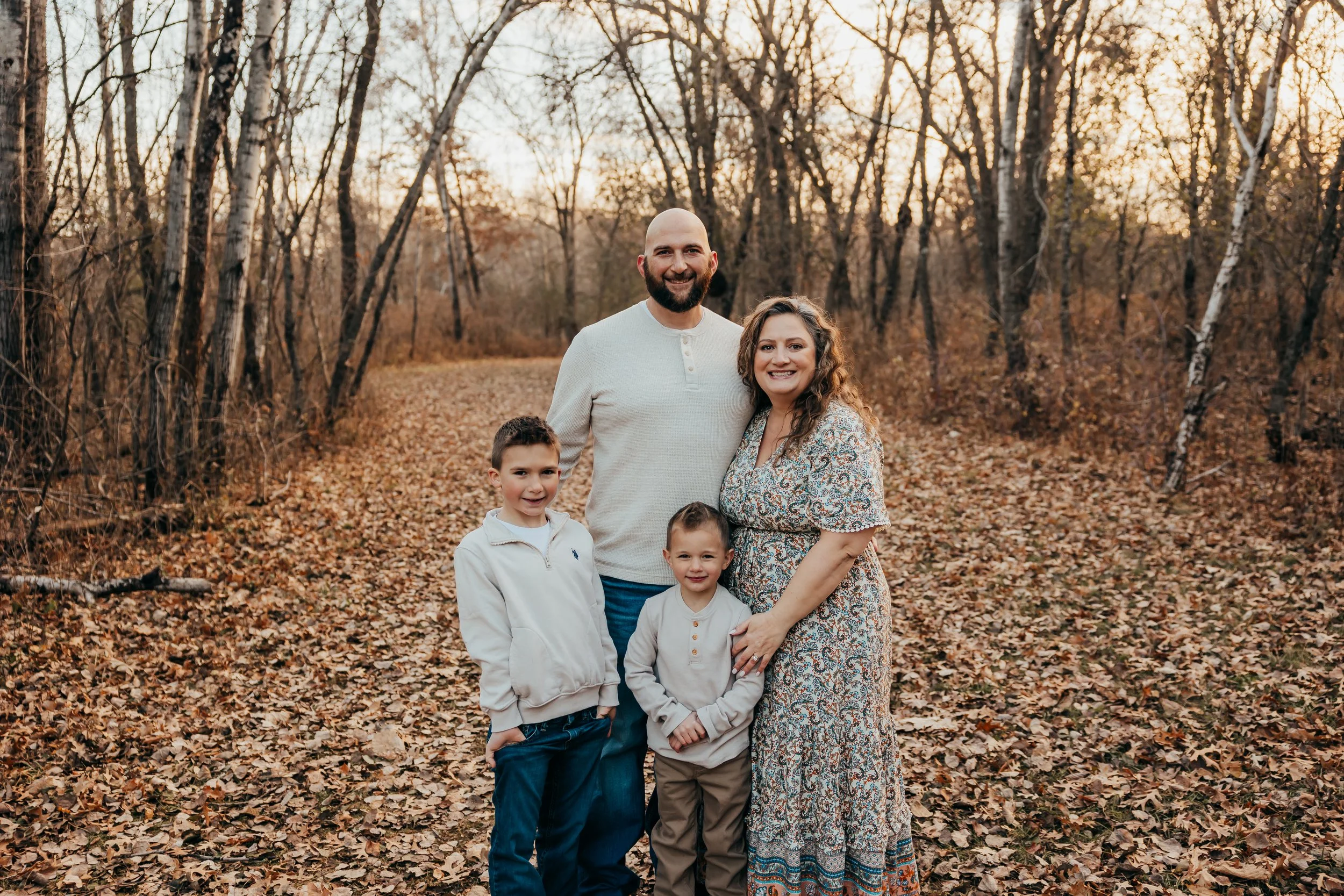 Family of four standing on a leaf-covered trail in a wooded area during autumn, smiling at the camera.