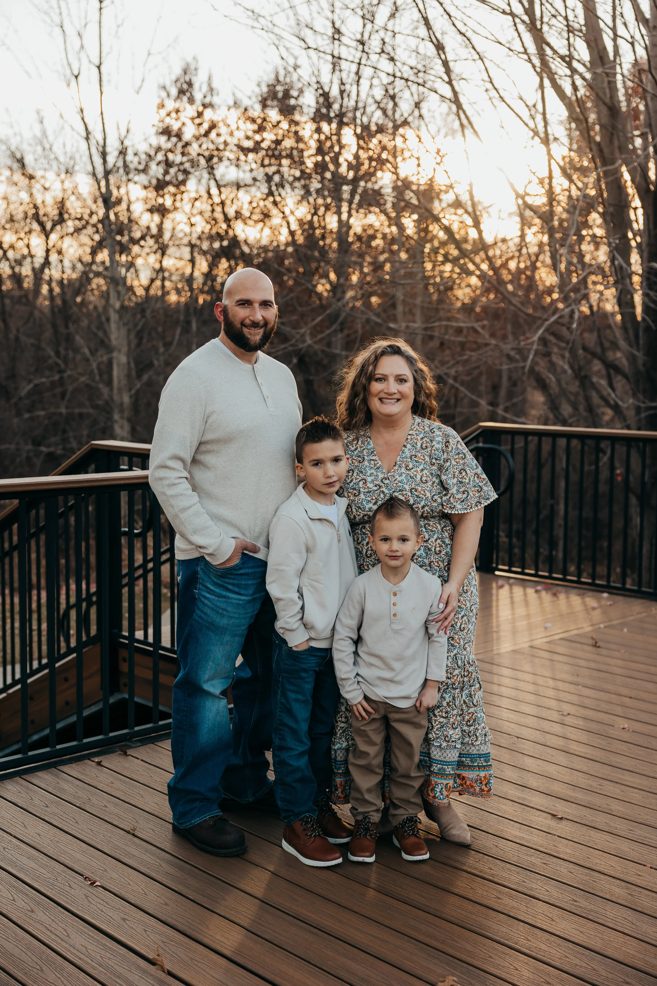 A family of five standing on a wooden deck outdoors during sunset. The background features leafless trees and a warm sky.